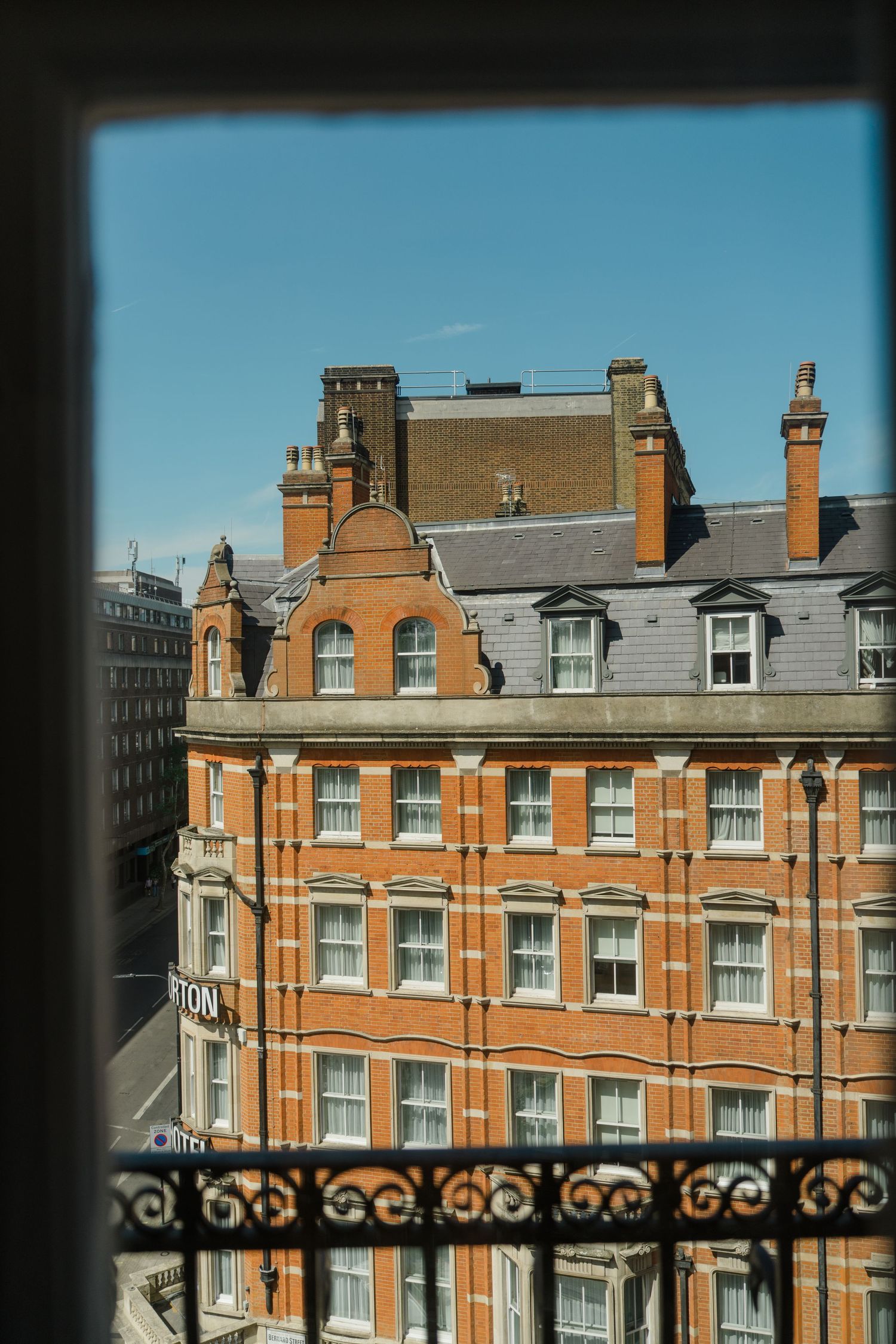 View through ornate iron balcony railing of grand Victorian red brick building against clear blue sky in London.