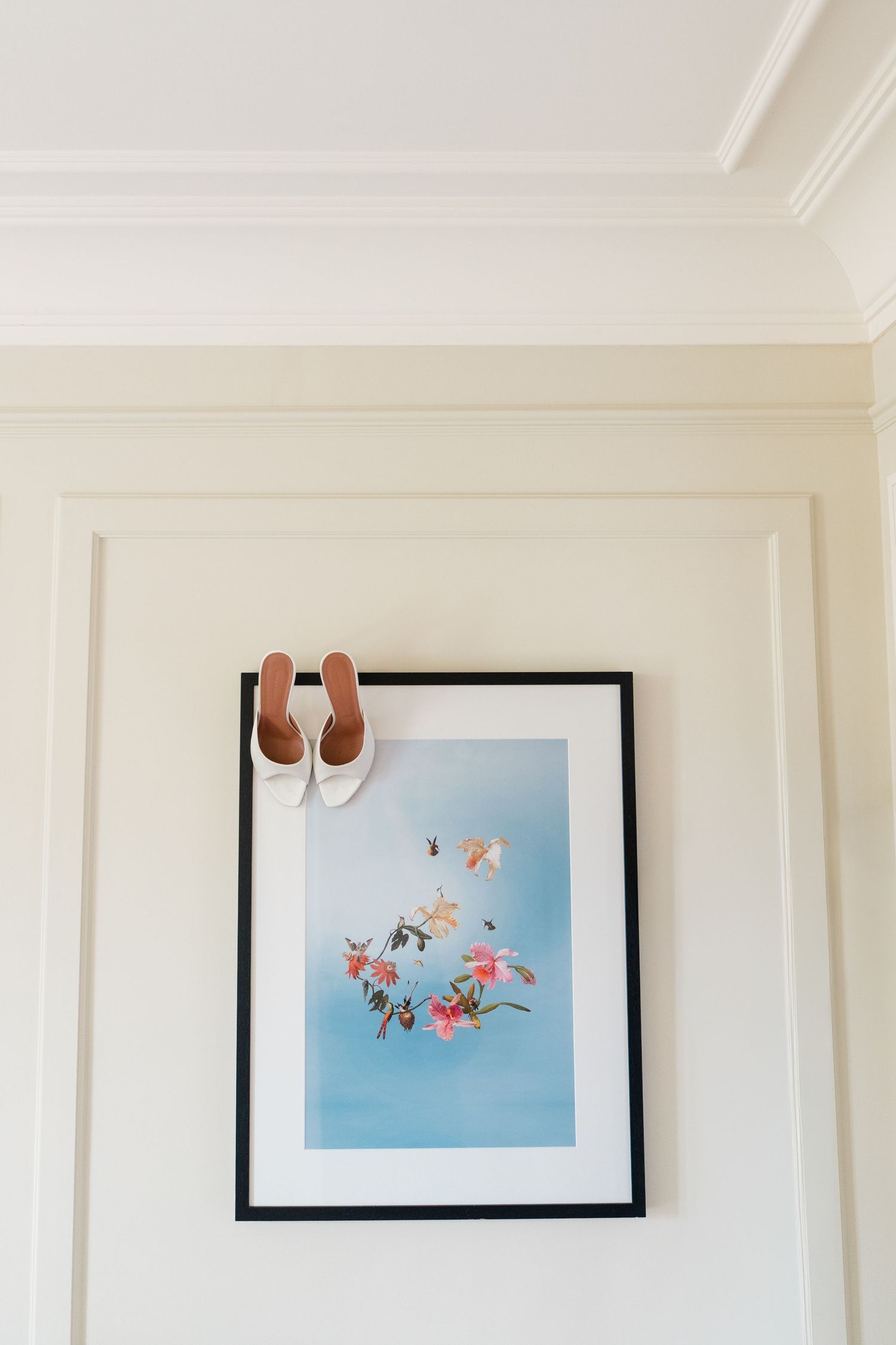 White bridal heels resting on a framed floral art print with blue background hanging on a cream wall.