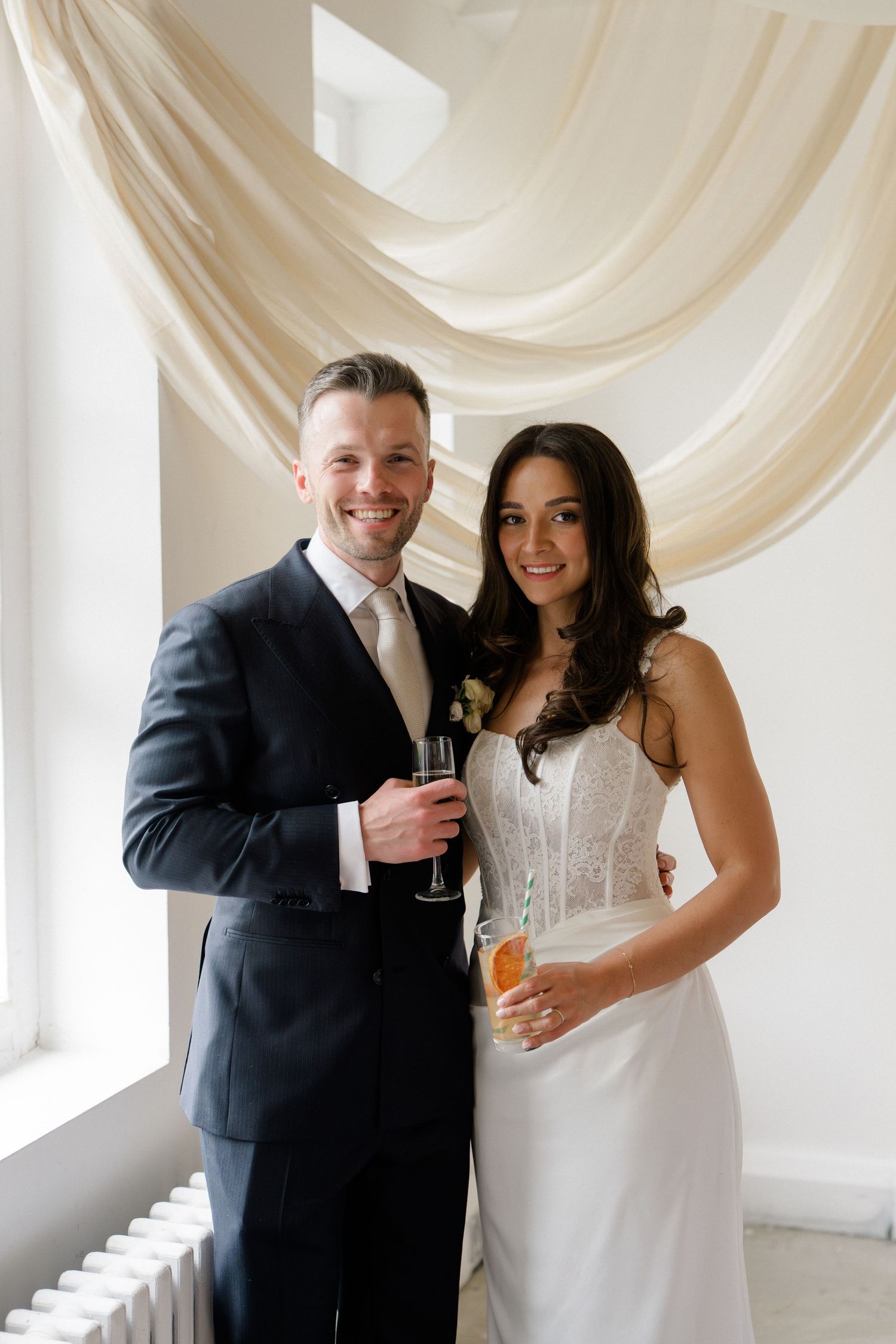 Smiling bride and groom holding champagne glasses posing together at wedding reception with draped fabric backdrop.
