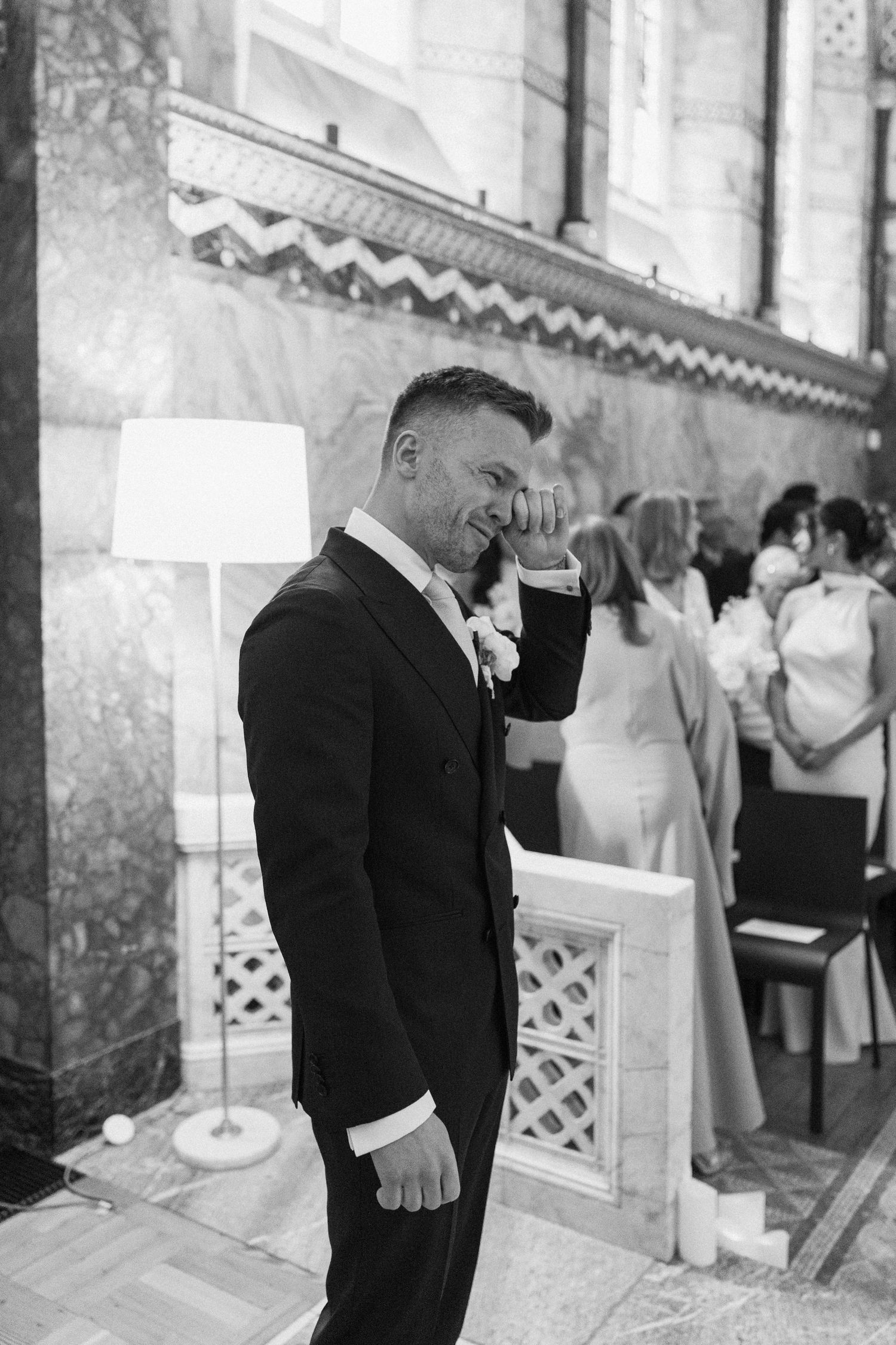 Emotional groom in black suit wiping tears during wedding ceremony in grand marble venue.