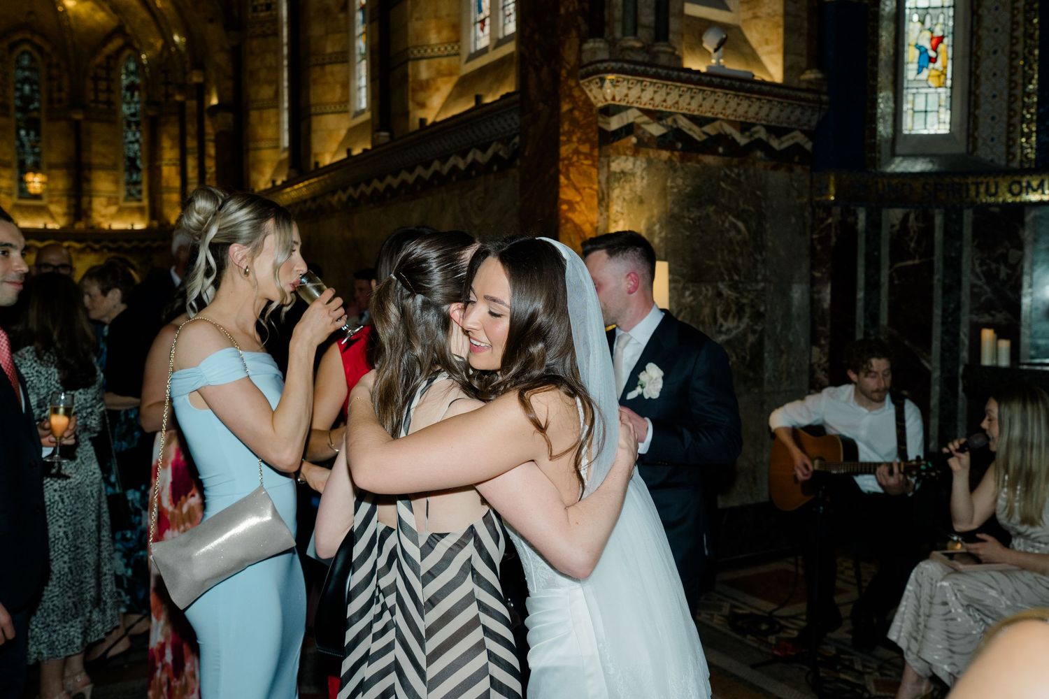 Bride in white gown and veil shares a warm hug with guests inside a decorated church.