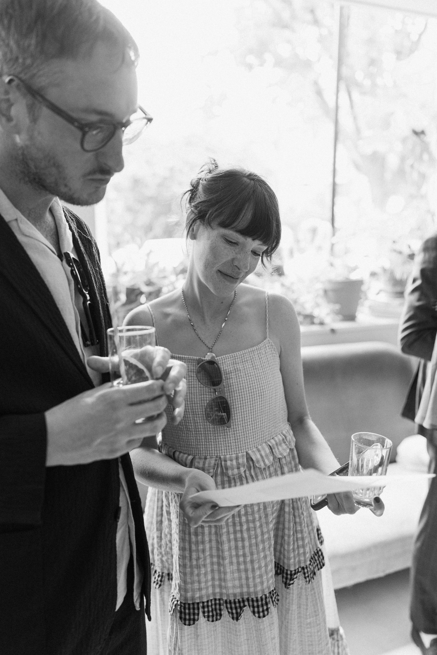 Wedding guests in black and white photo review ceremony program while holding drinks at reception.