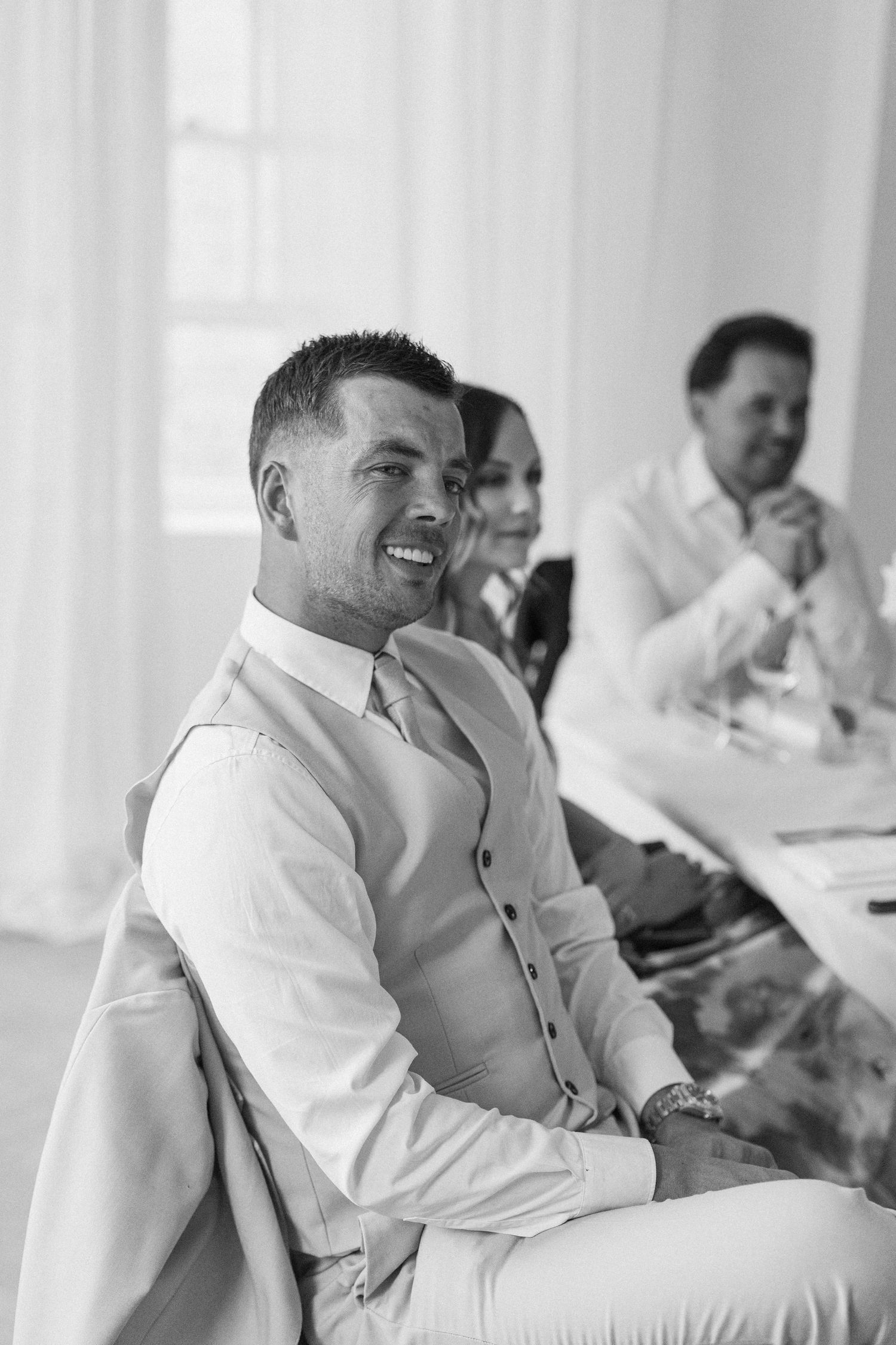 Smiling groom in white vest seated at wedding reception table with guests in background.