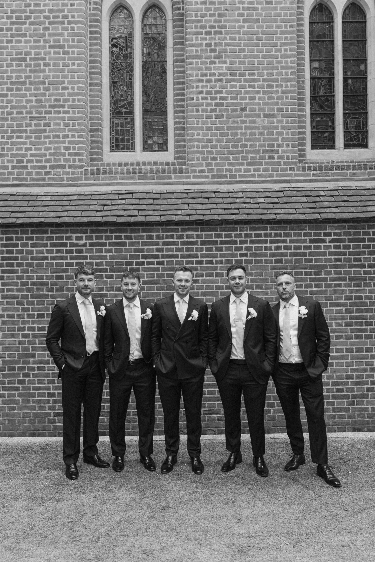 Five groomsmen in black suits with boutonnieres pose in front of a brick church wall in black and white.