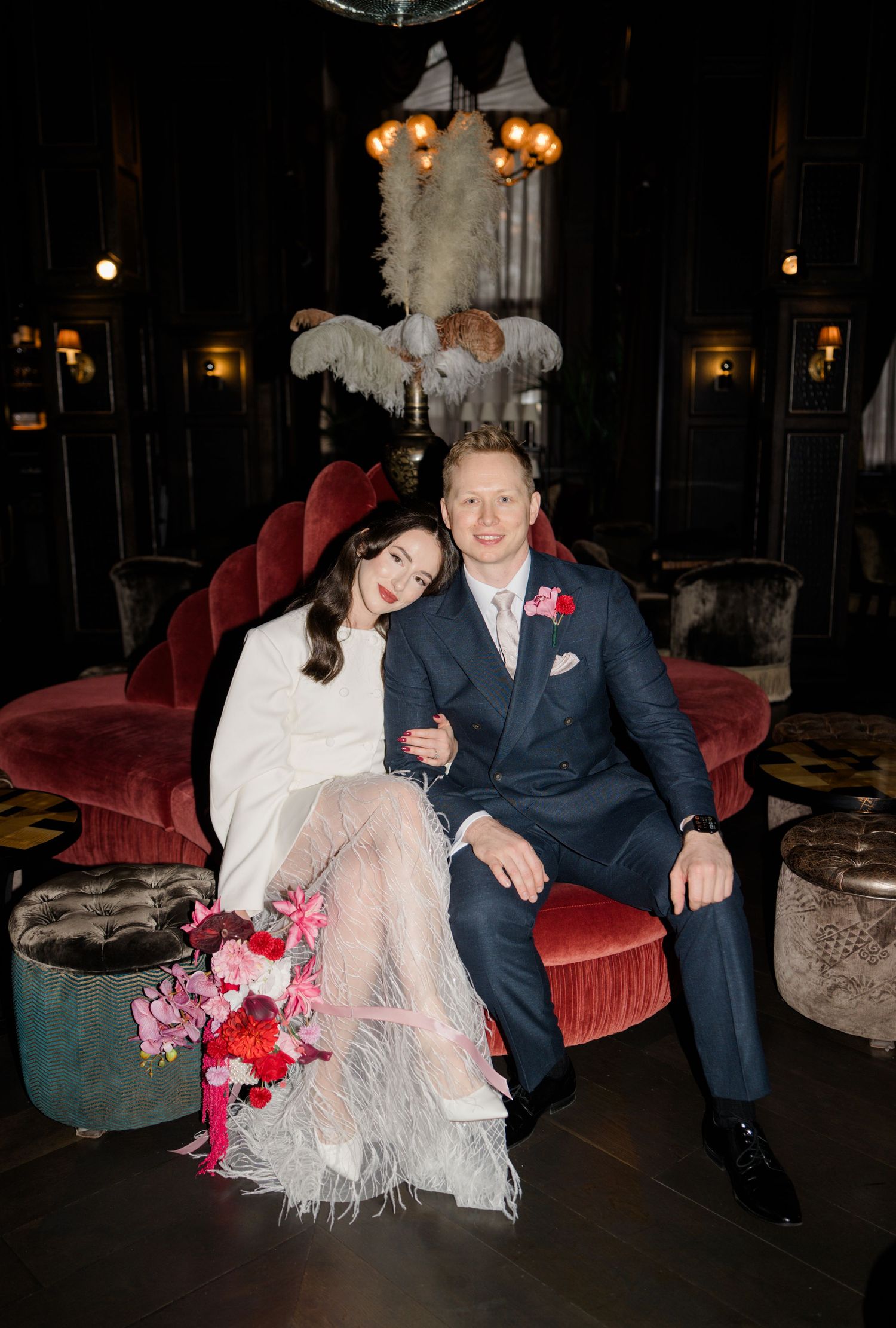 Bride and groom seated on velvet sofa in dark luxe venue, bride holding pink floral bouquet.