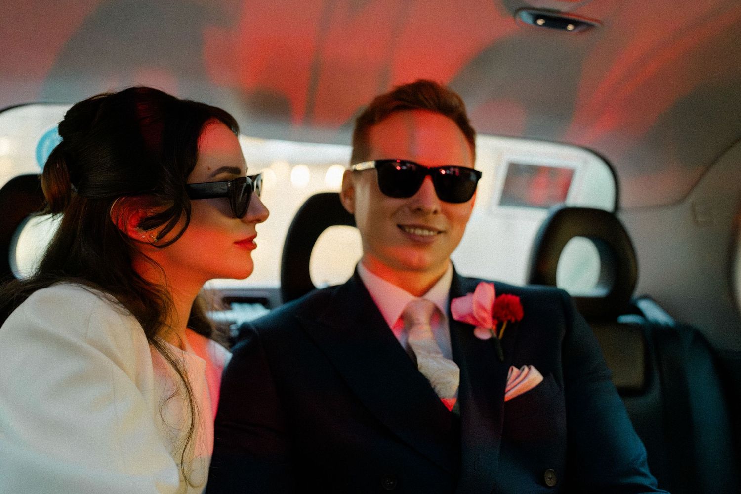 Bride and groom wearing sunglasses smiling together in car with pink boutonniere wedding detail.
