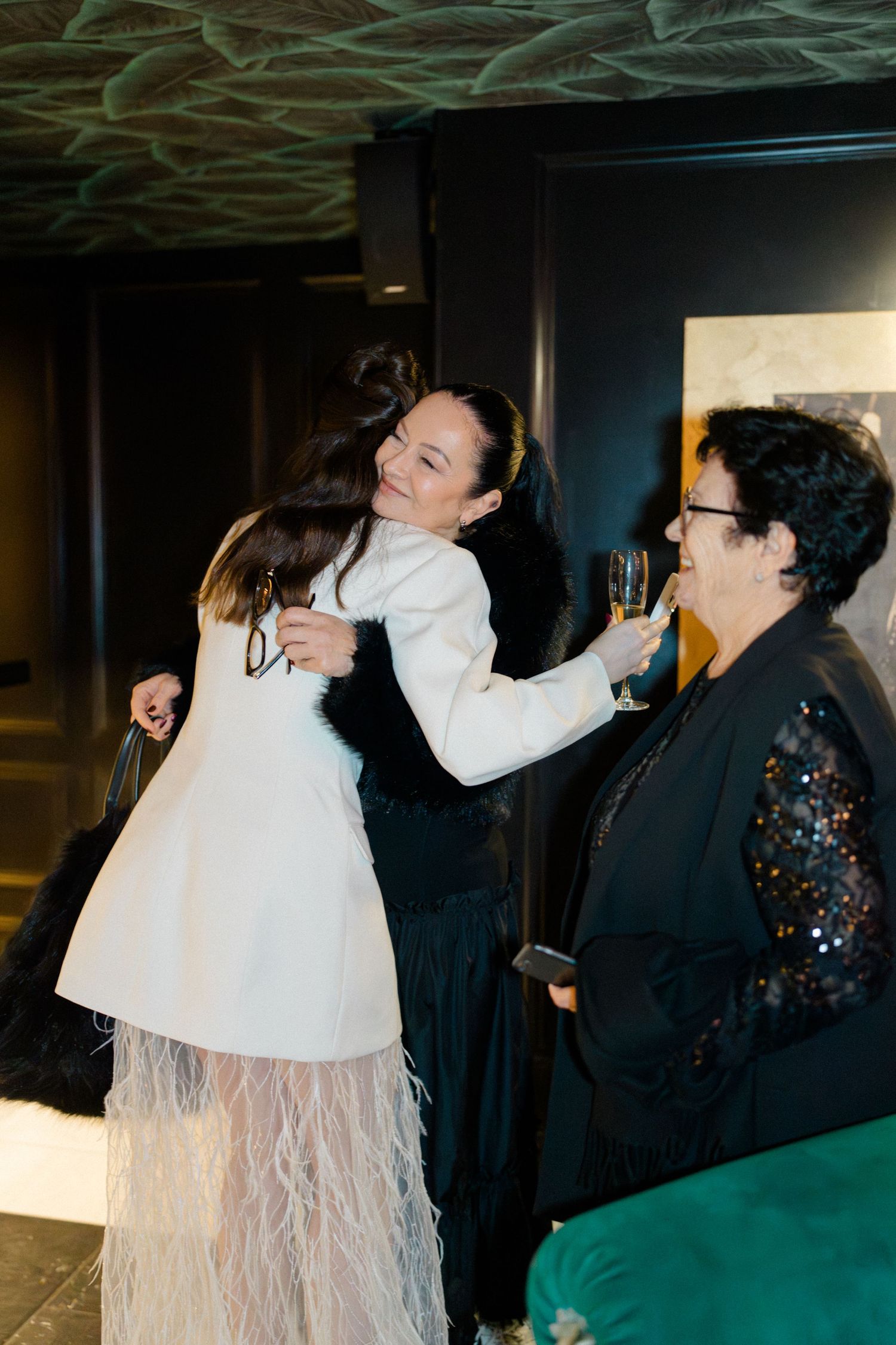 Bride in white feather hem blazer dress embraces a guest while another woman in sequins looks on at wedding reception.