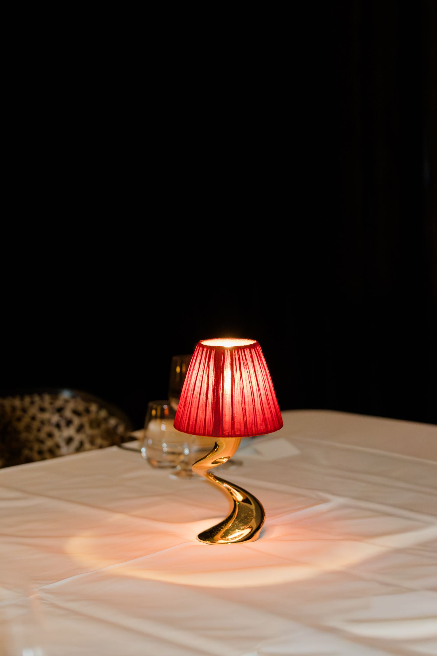 Elegant red lamp with gold twisted base glows warmly on a white tablecloth in a dark restaurant.
