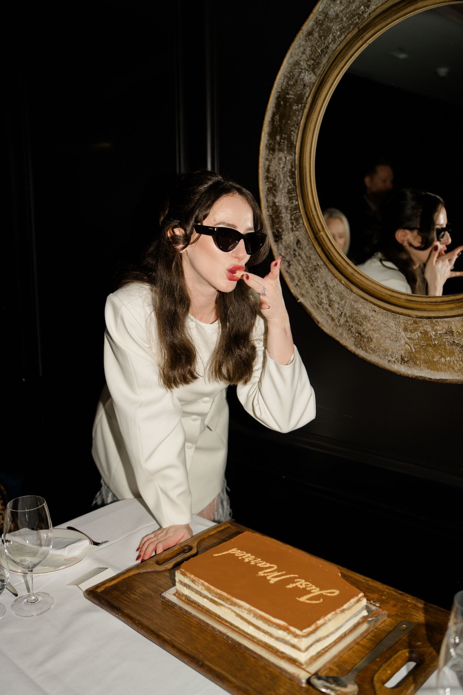 Bride in sunglasses and white blazer tastes wedding cake labeled Just Married in front of a round vintage mirror.