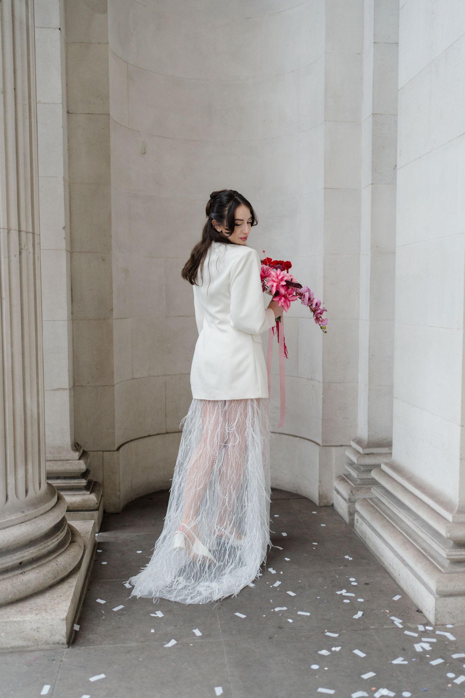 Bride in white blazer dress with feather skirt holds pink bouquet against stone columns.