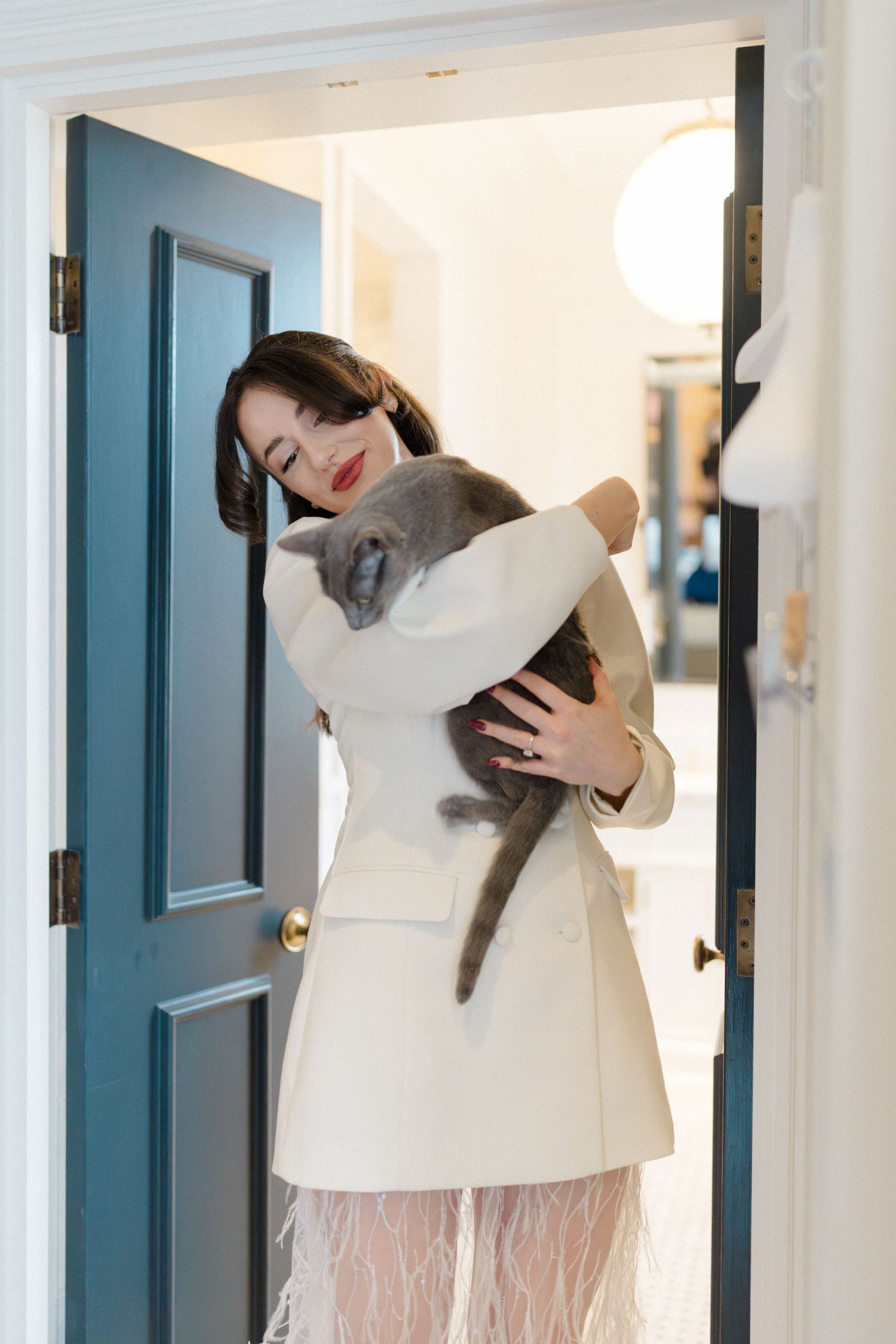 Beautiful bride in white blazer and feathered skirt lovingly holds a grey cat in a doorway.