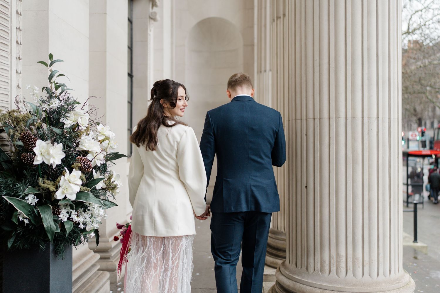 Bride and groom hold hands walking past large floral arrangement between stone columns.