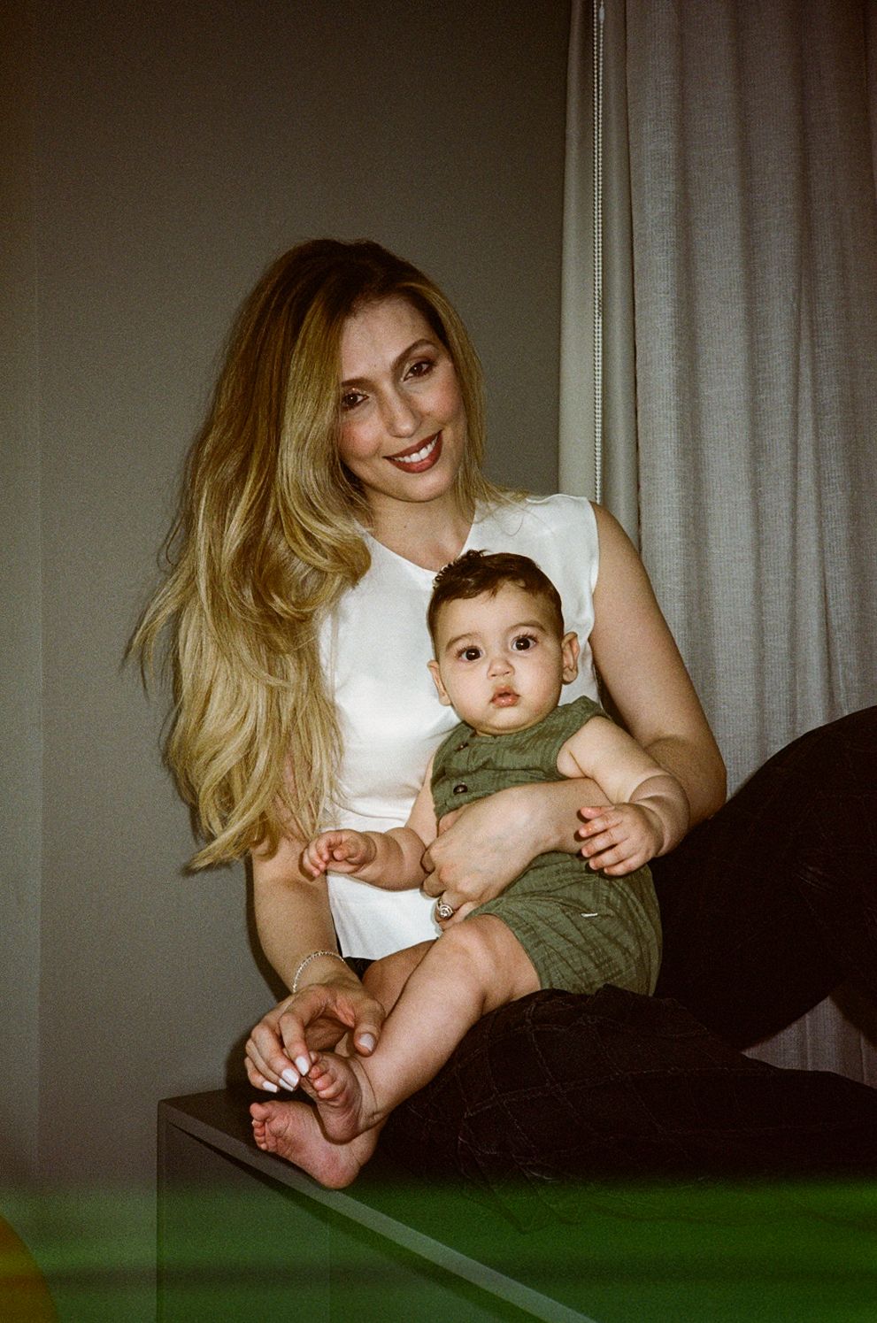 Smiling blonde woman holding a baby in a green outfit, sitting together in a warmly lit room.