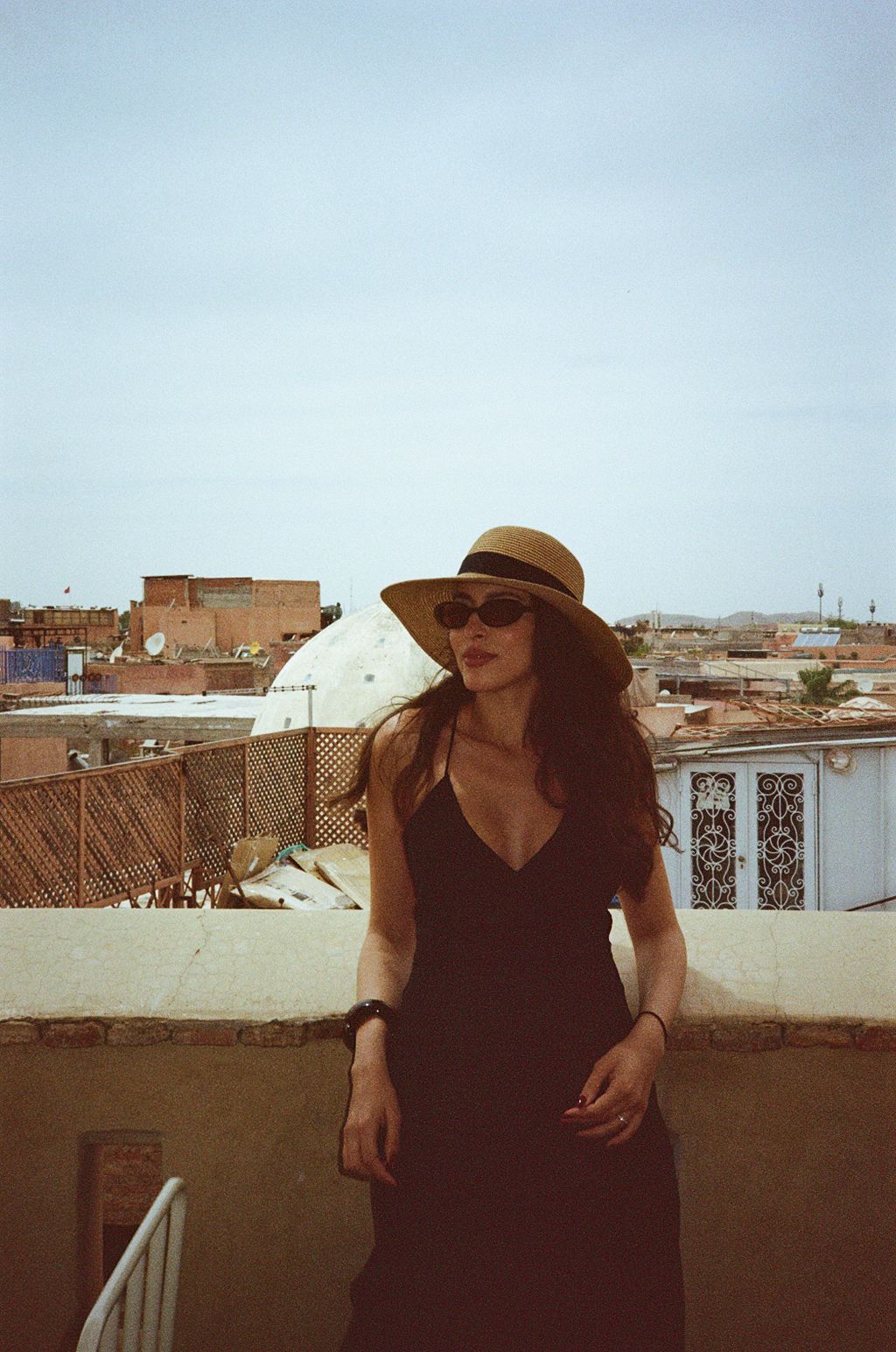 Woman in black dress and straw hat standing on a rooftop with a city skyline in the background.
