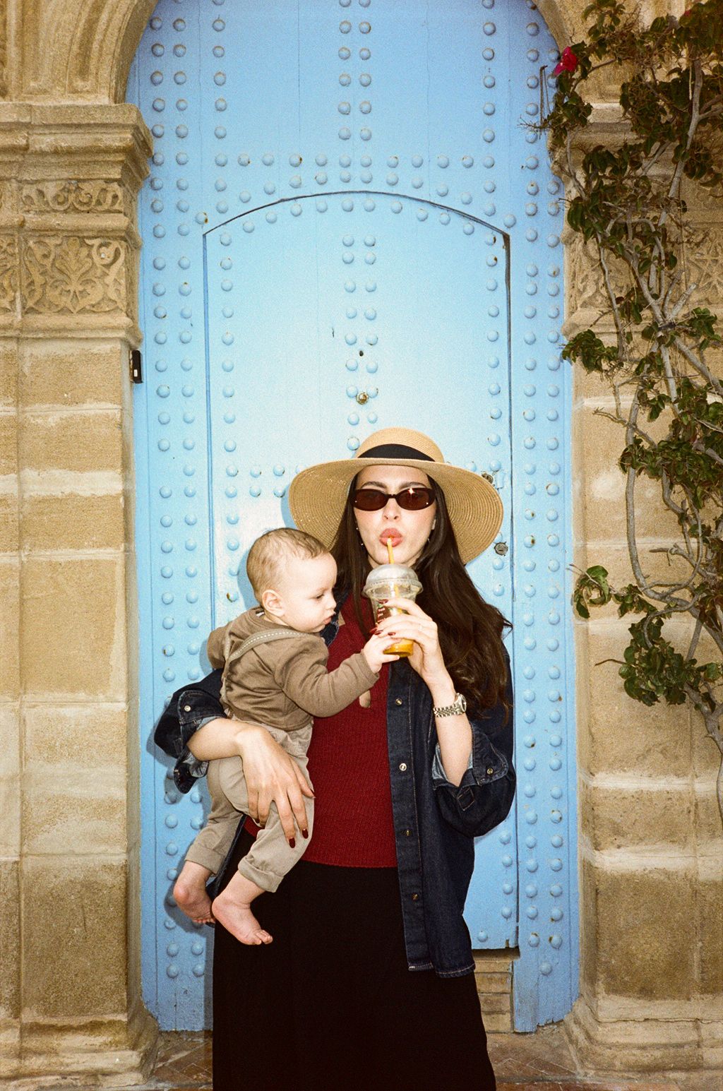 Woman in straw hat and sunglasses holding baby while drinking from cup in front of blue studded door.