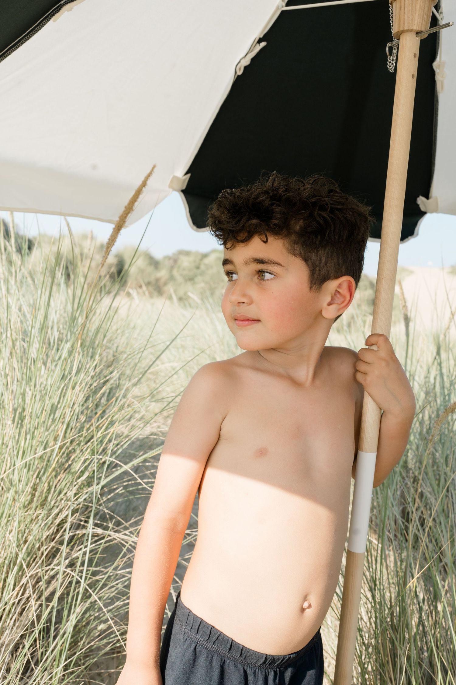 Child under white beach umbrella surrounded by sand dunes and beach grass.