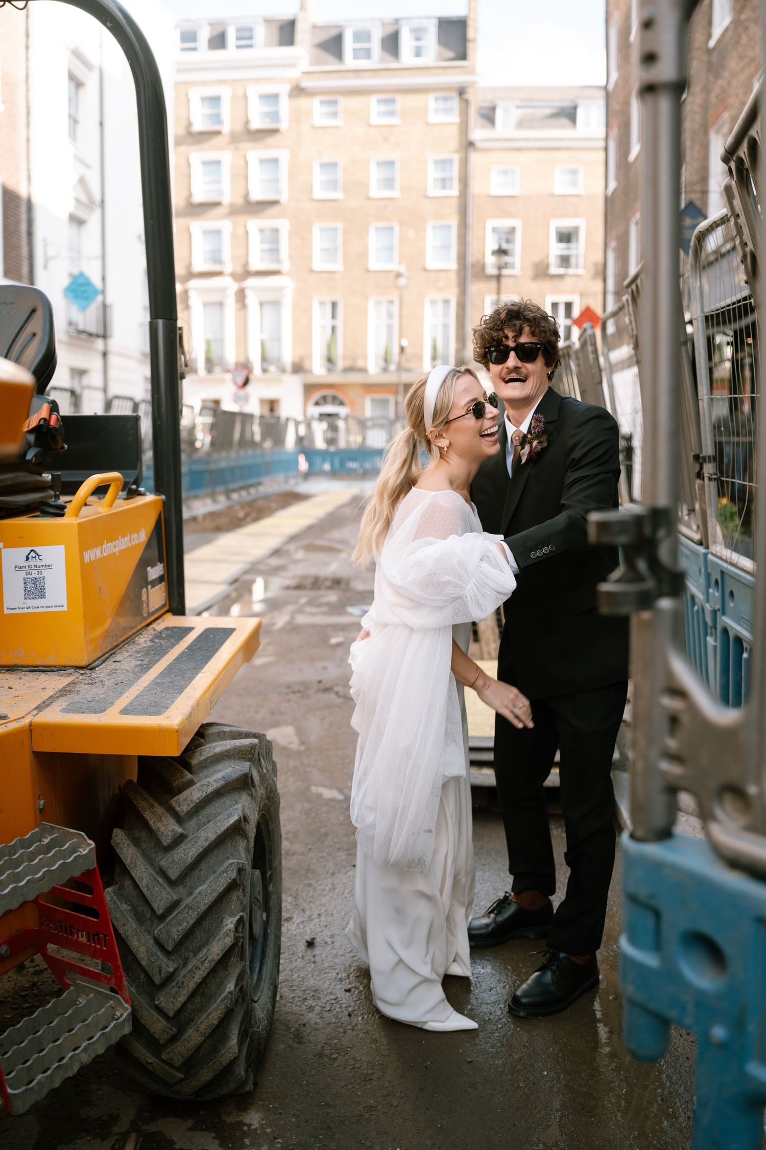 A couple in wedding attire poses playfully next to a yellow construction vehicle on an urban street.