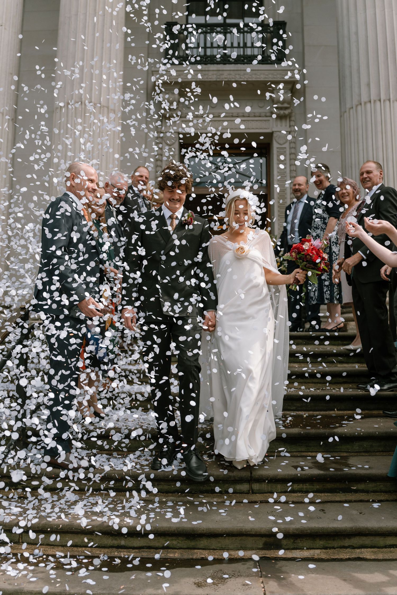 Shower of white confetti fills the air during wedding celebration on stone steps.