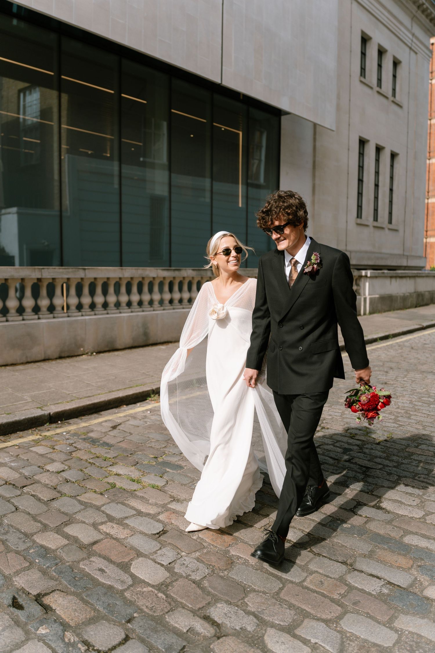 A couple strolls along cobblestone streets holding red flowers against urban architecture.