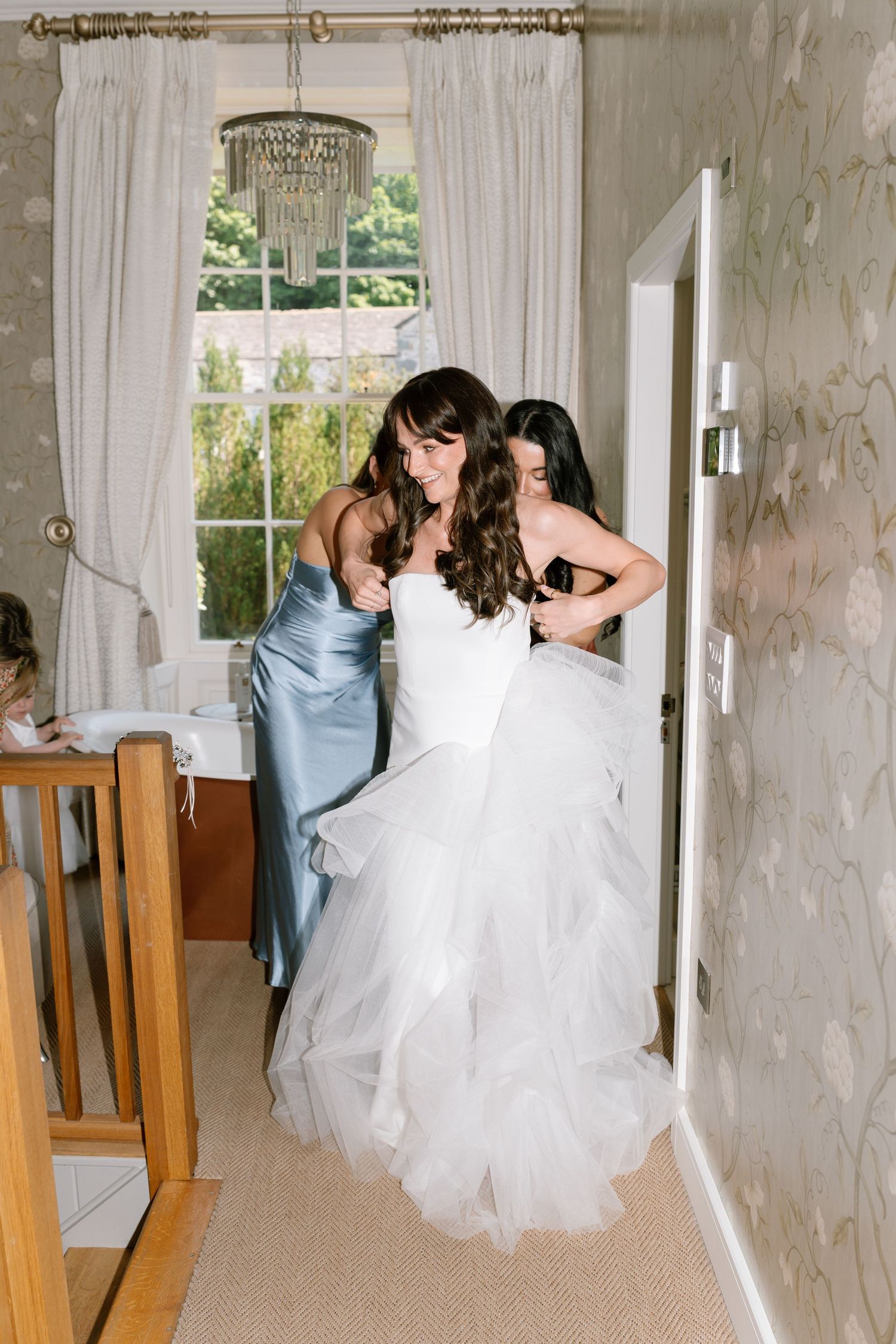 A bride in a white wedding gown gets ready in an elegant room with floral wallpaper and vintage decor.