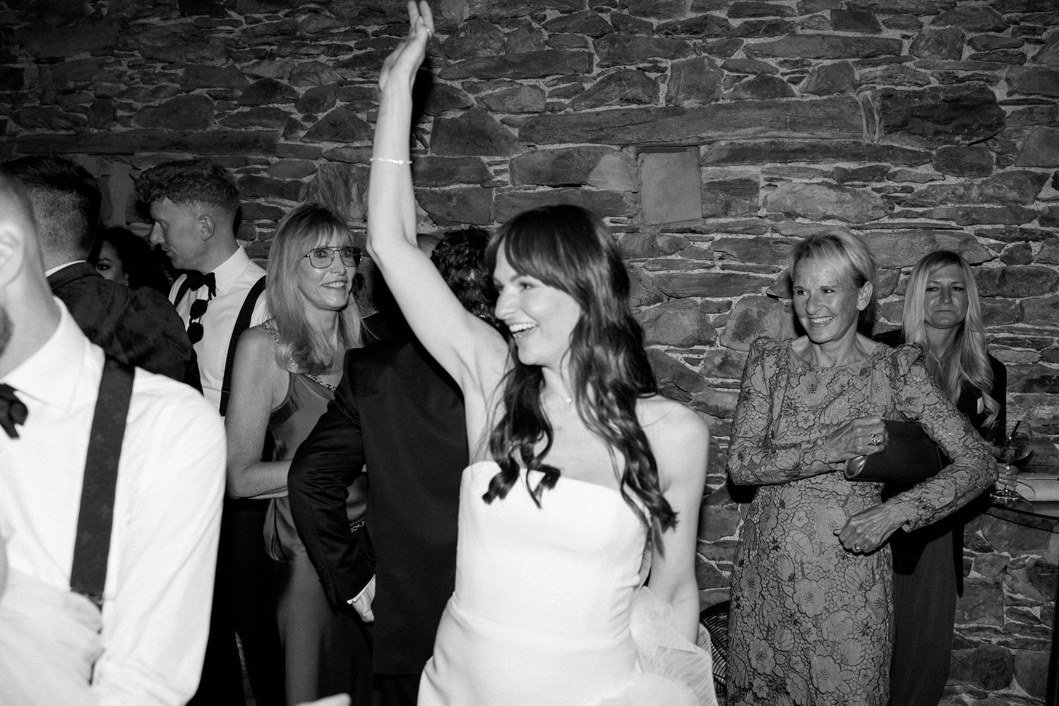 A bride in white raises her hand while dancing at a wedding reception with stone walls.