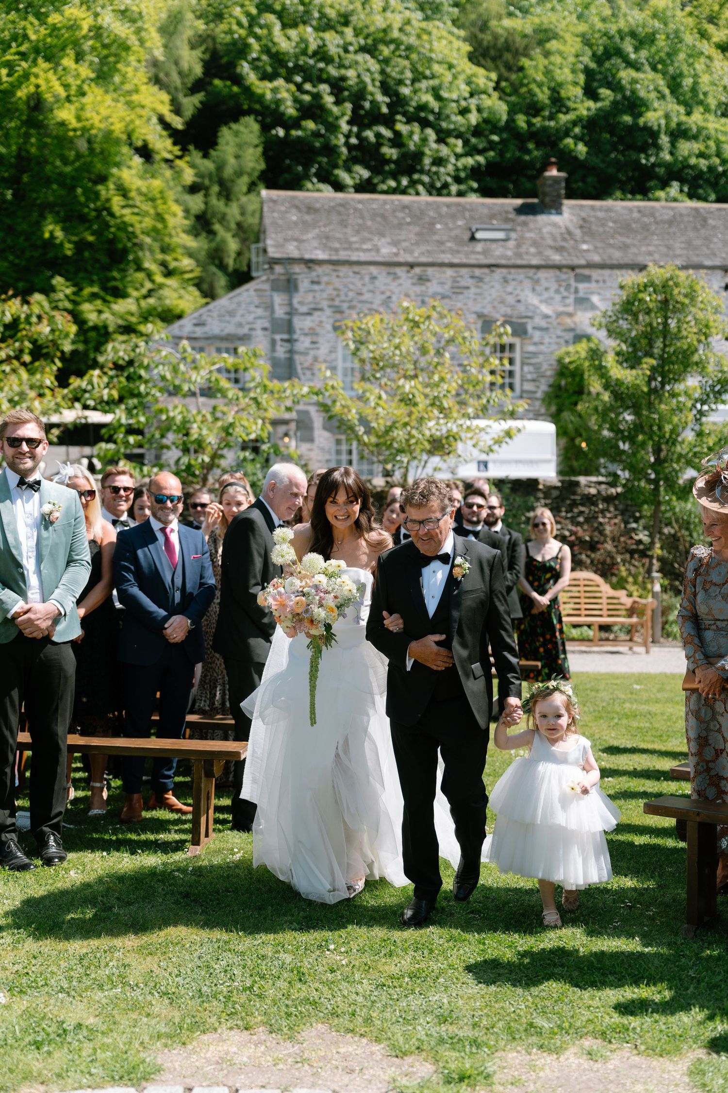 Wedding party poses for photos on a sunny lawn with stone building backdrop.