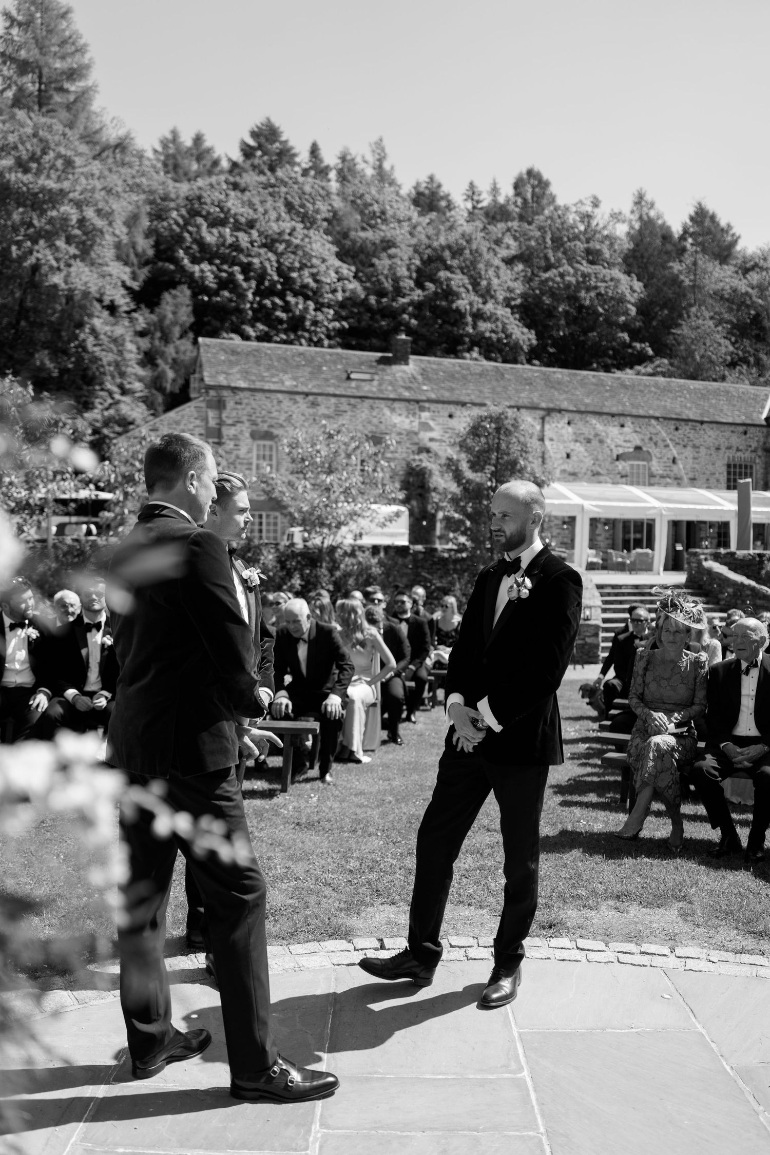 Black and white photo of formally dressed guests standing on a marked outdoor area.