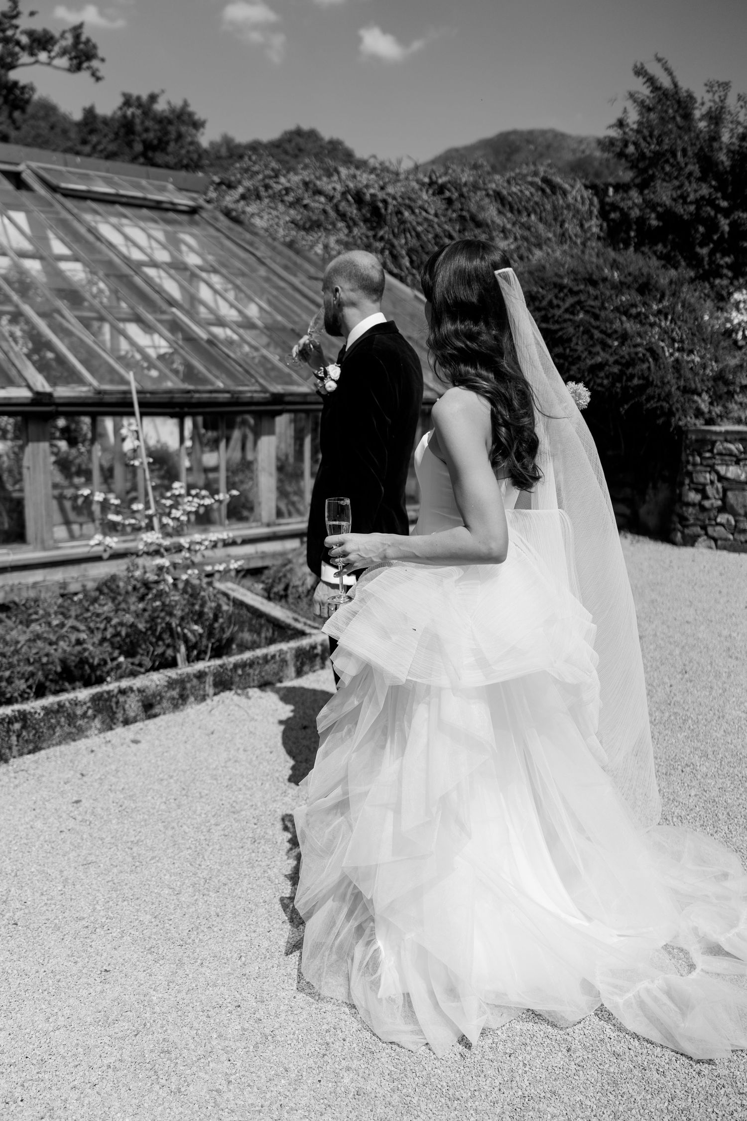 A black and white photo of a couple near greenhouse windows in a garden setting.