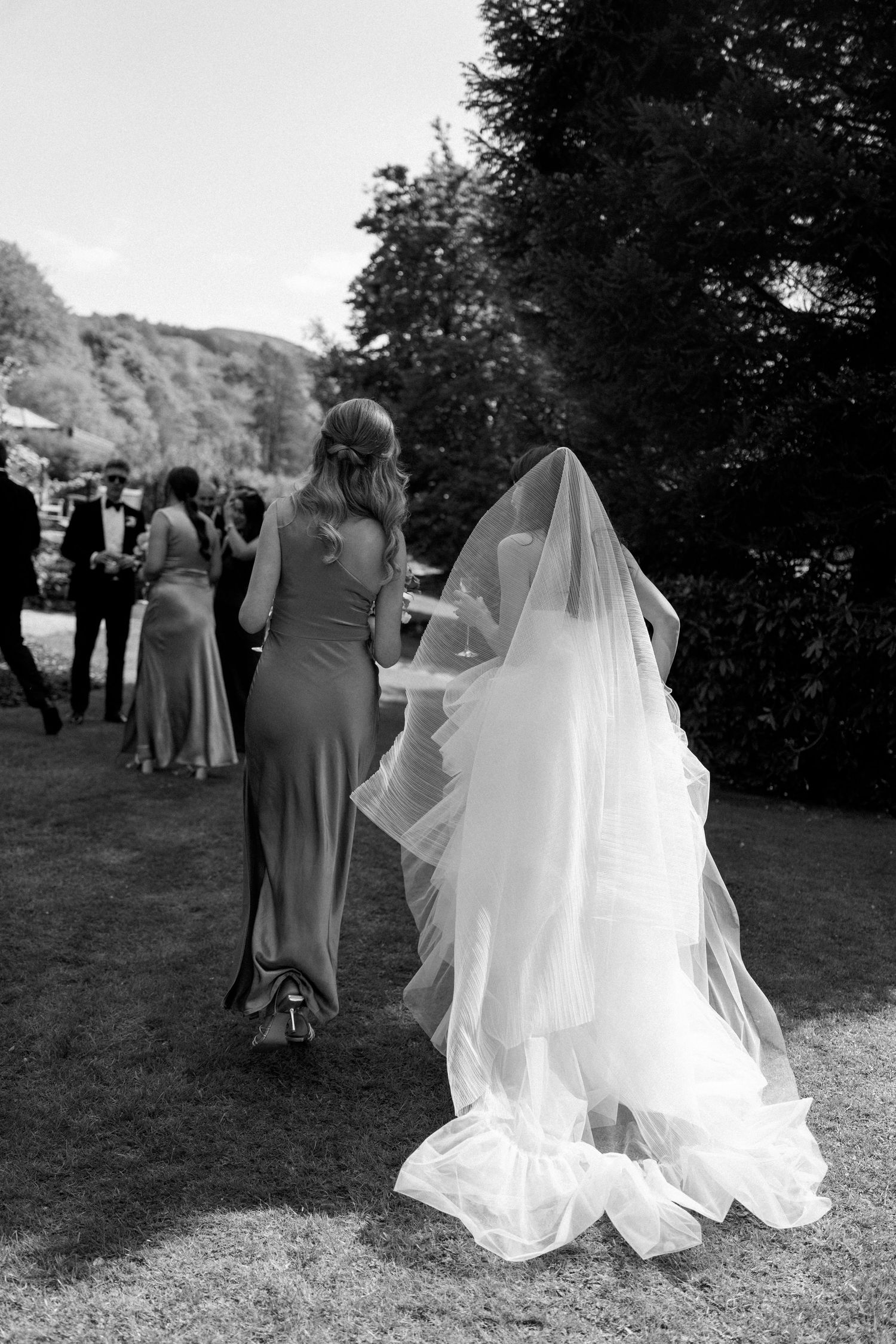Bride in flowing white dress walks with wedding party across a scenic garden lawn.