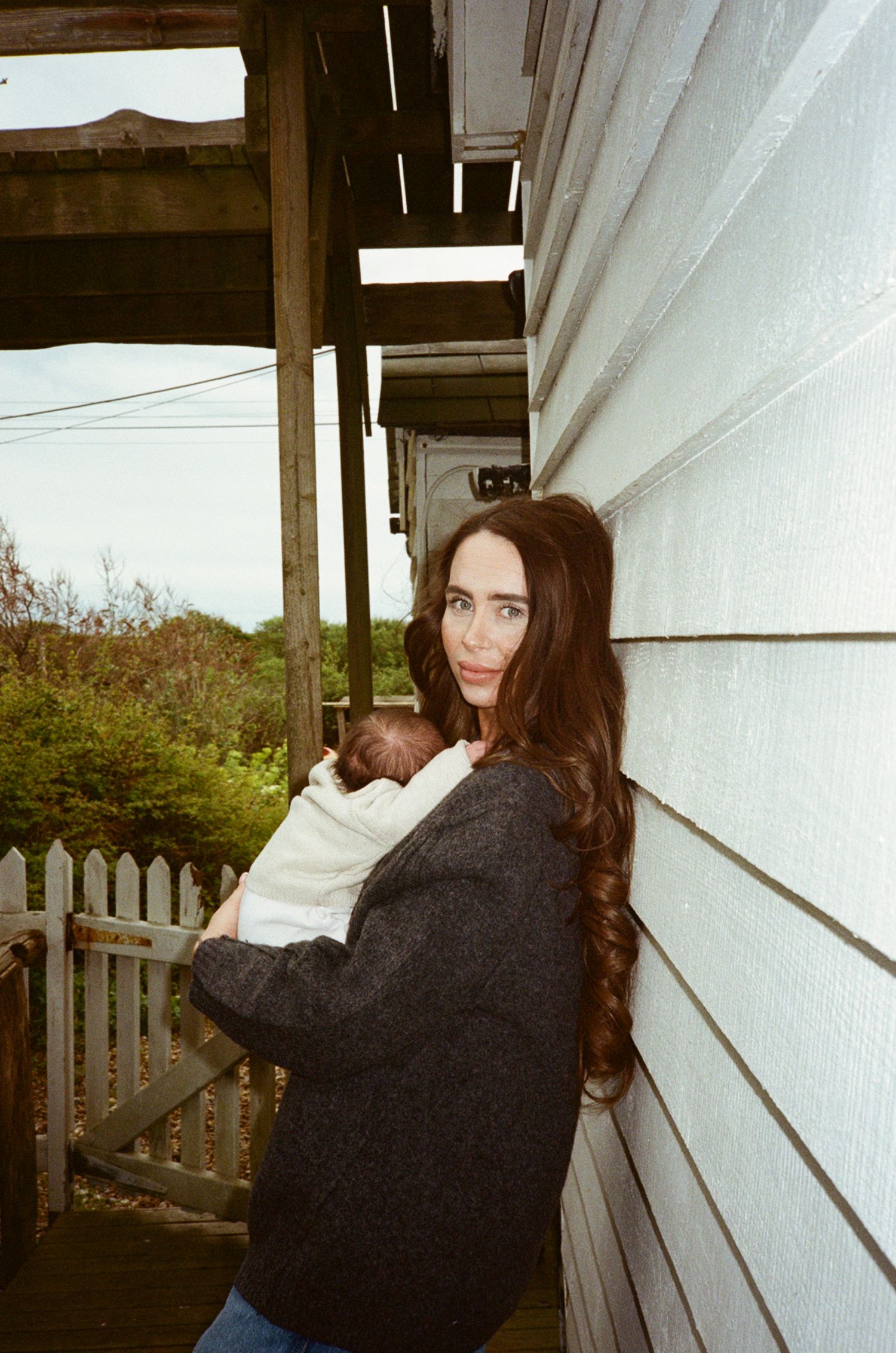 A woman with long dark hair holds a newborn baby on a porch, leaning against a white clapboard house.