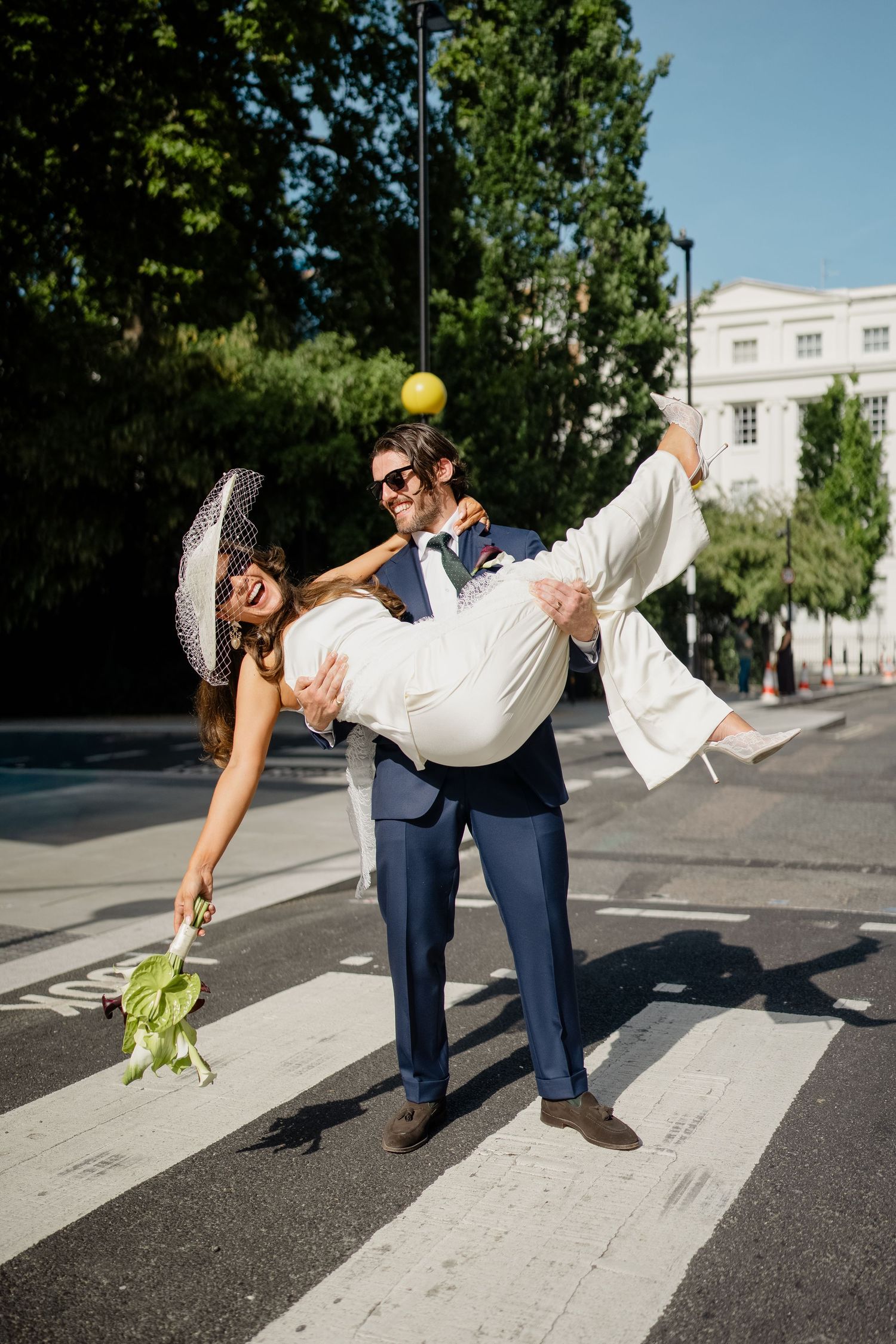 Groom carries laughing bride across zebra crossing while green apple flies through the air.