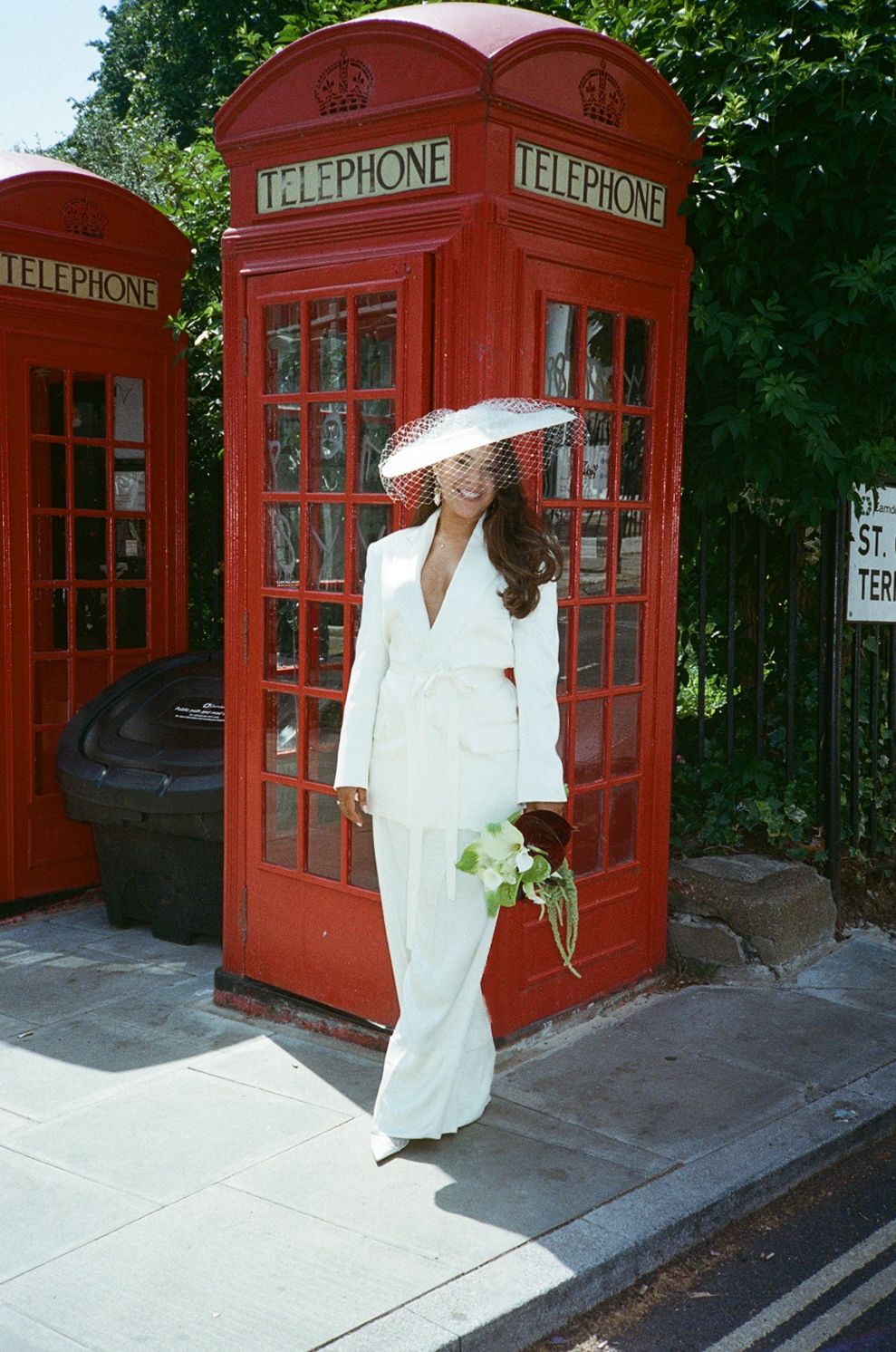 Bride in white suit and hat poses by iconic red London telephone boxes holding a bouquet.