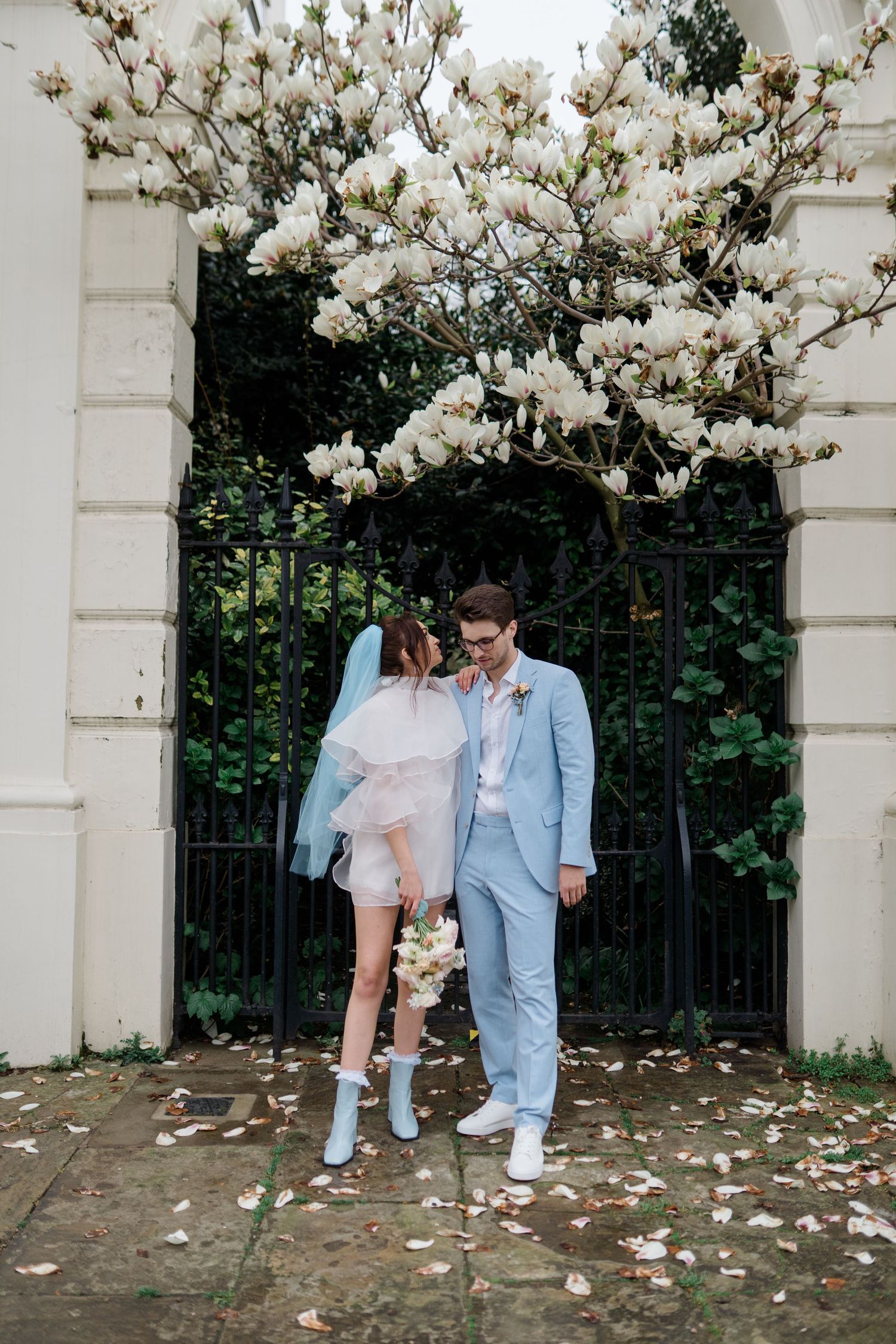Bride in short white dress with blue veil and groom in light blue suit stand under blooming magnolia tree by iron gate.