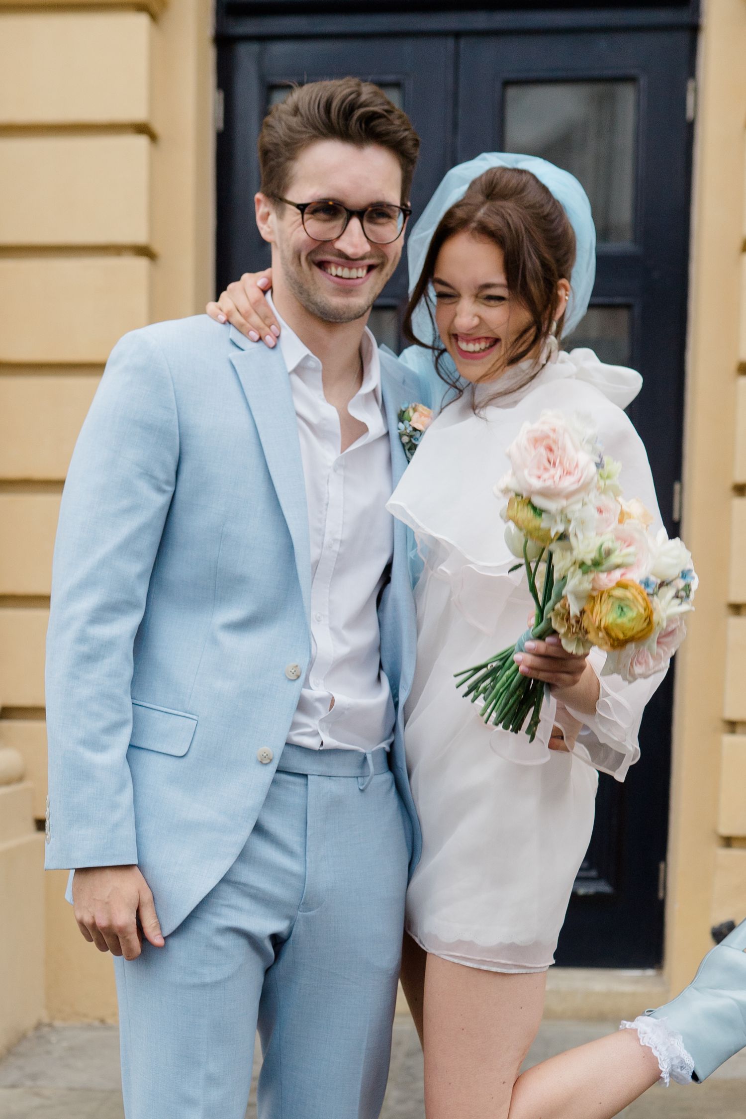 Happy newlyweds posing outdoors, groom in light blue suit and bride in white mini dress holding a floral bouquet.