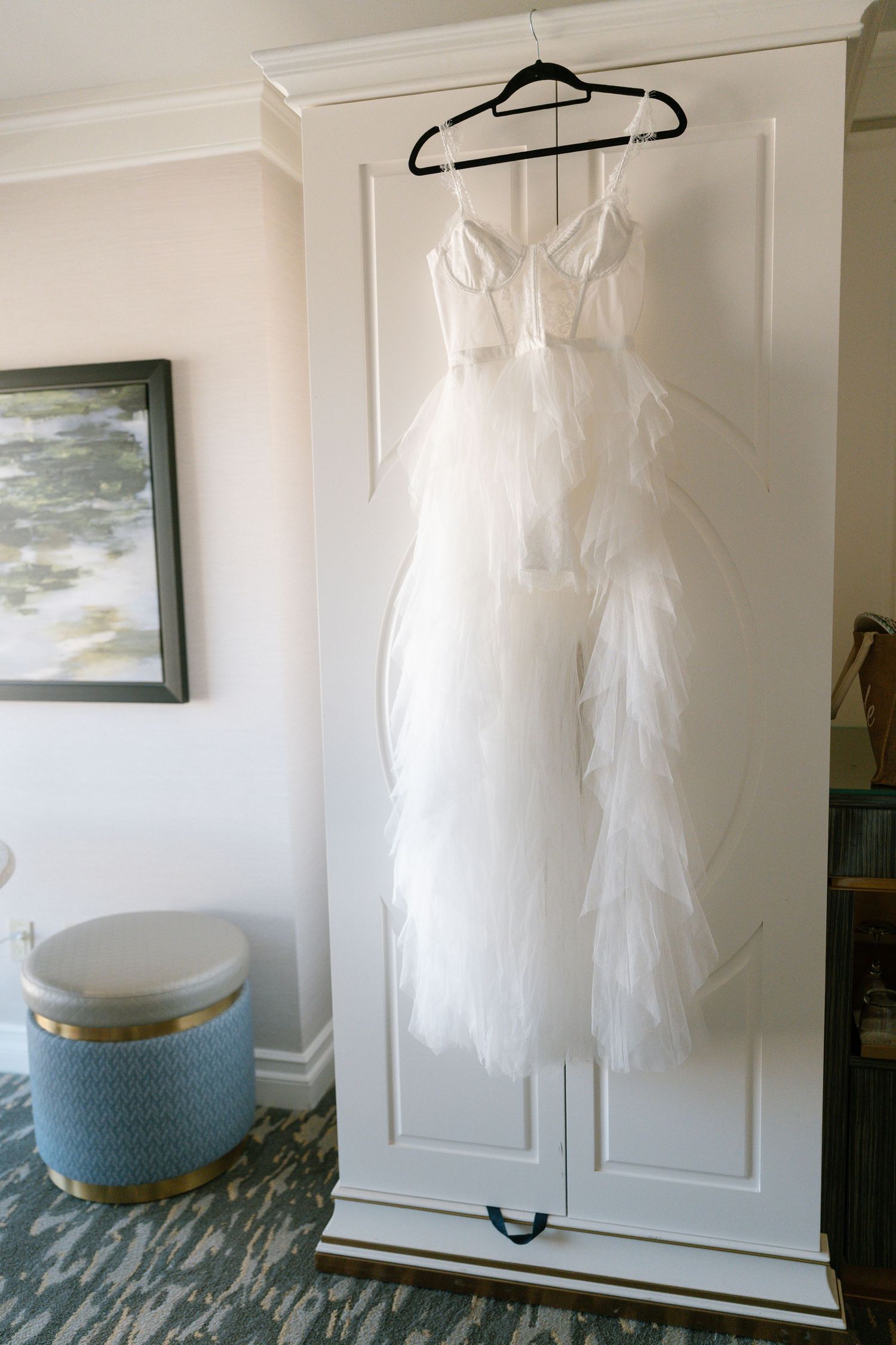 Elegant white wedding gown displayed on hanger against white wardrobe with blue ottoman nearby.