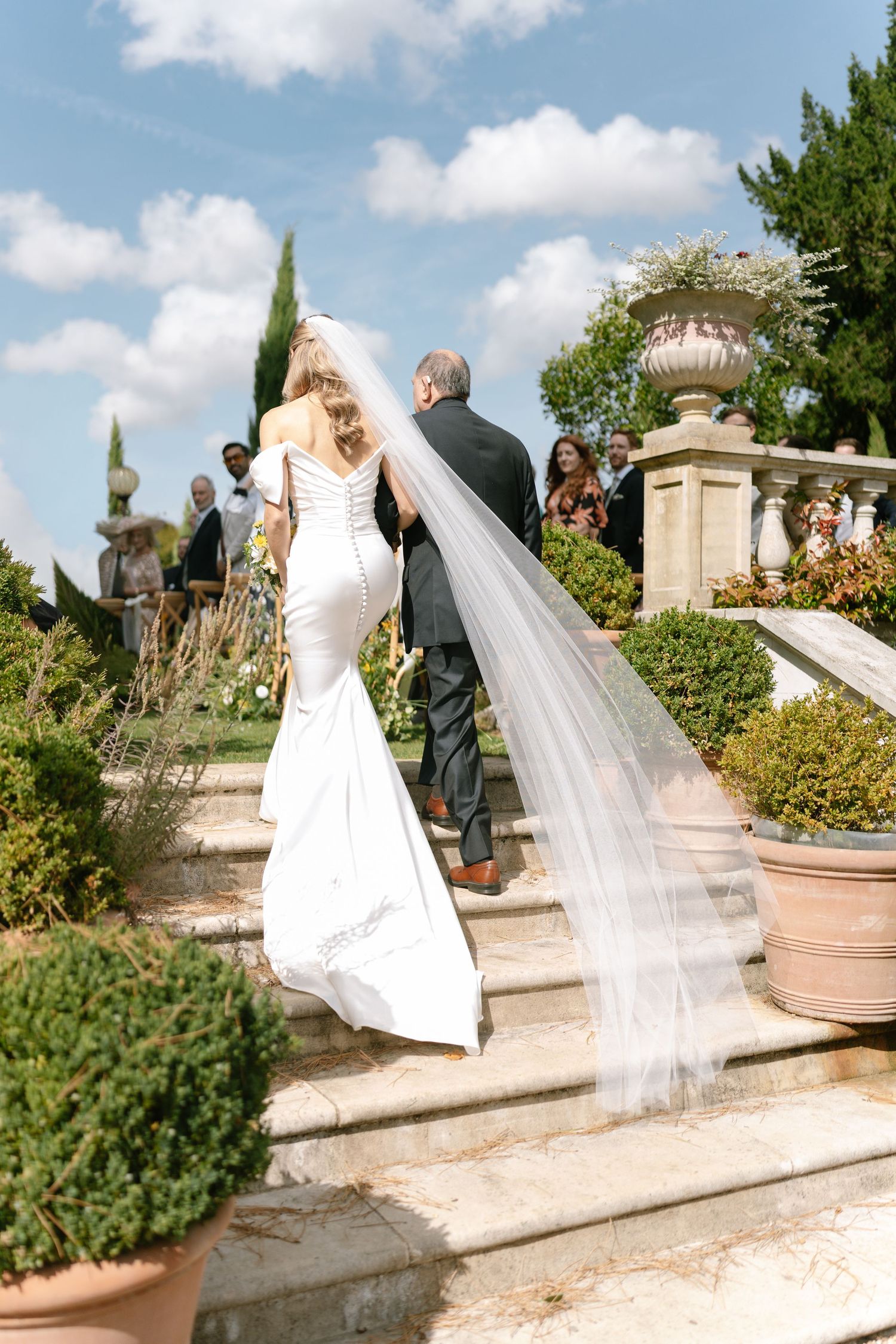 Wedding couple walking down stone steps with flowing veil in sunny garden setting.