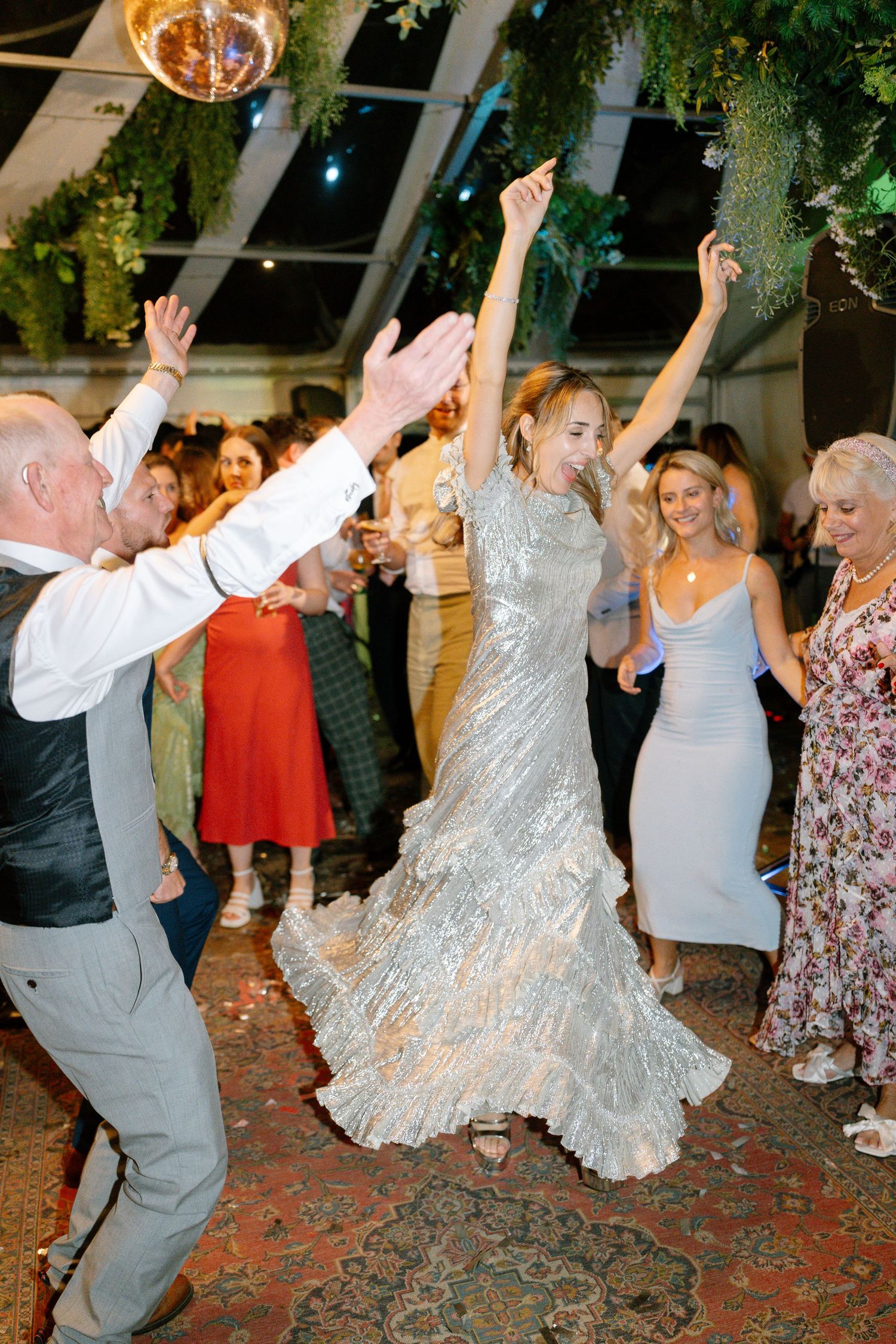 Wedding party dancing energetically on a patterned carpet at an indoor reception.