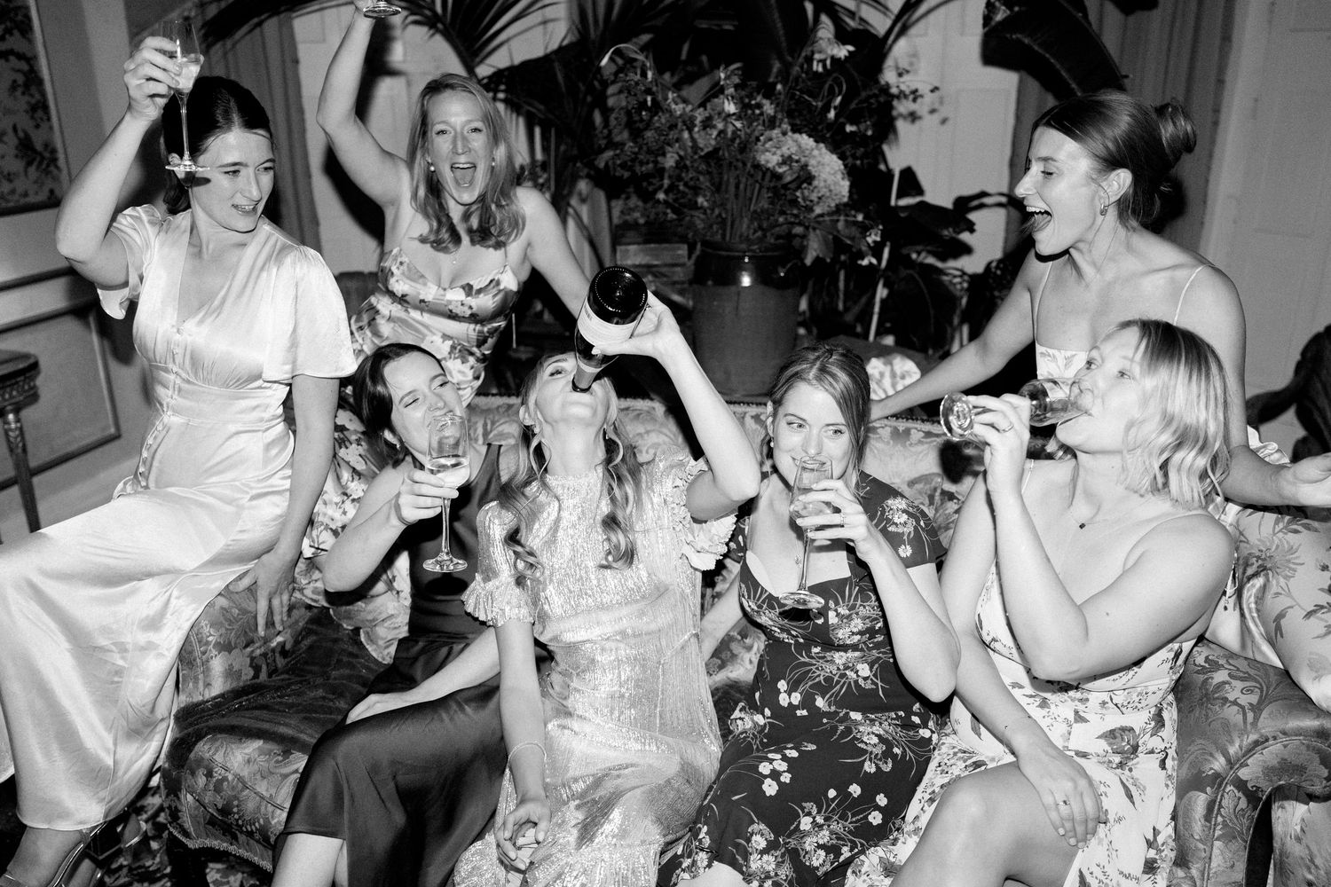 Wedding party laughing and drinking champagne while seated on elegant furniture.