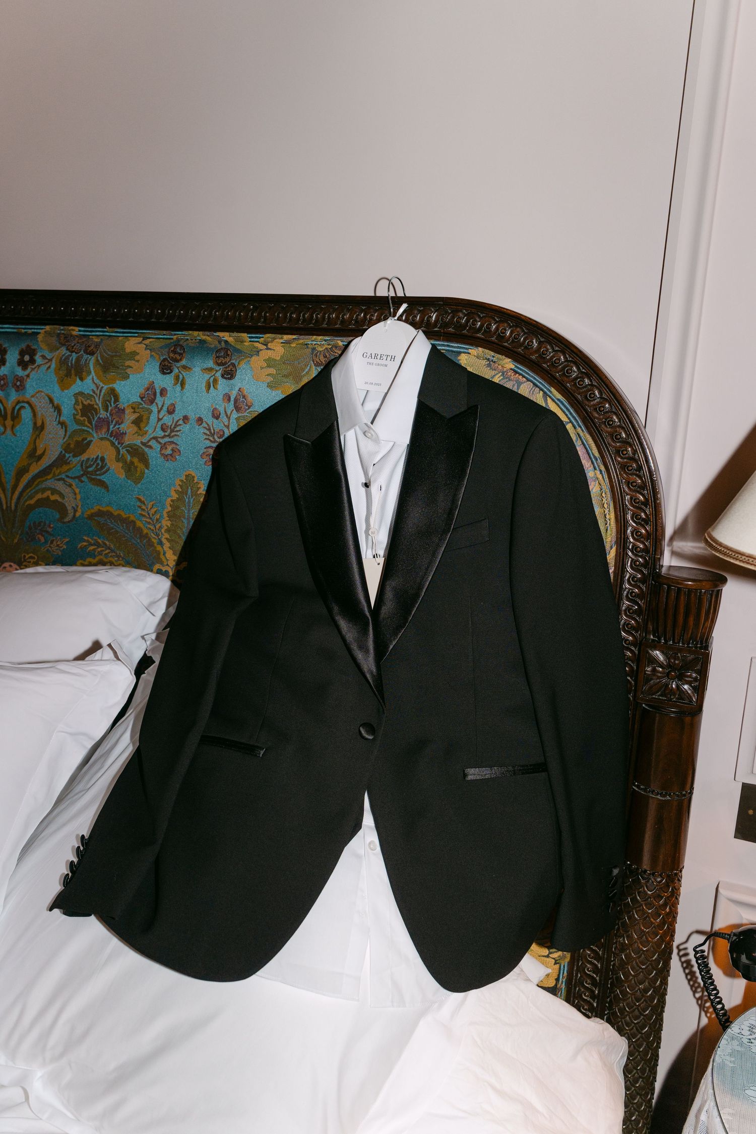 Black tuxedo jacket displayed on hanger against ornate vintage headboard.