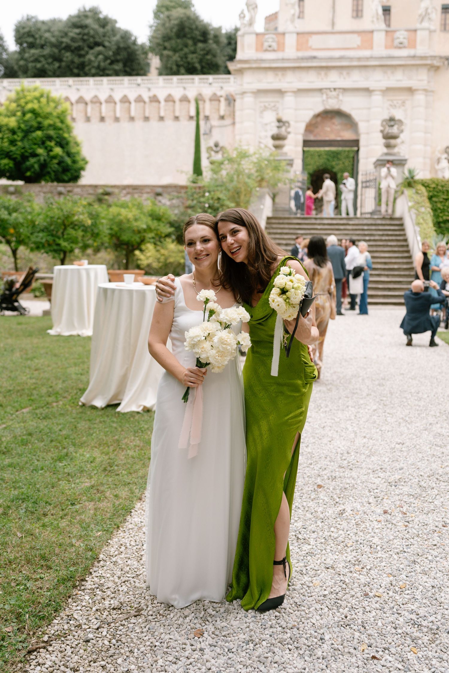 Two people in elegant attire pose with white bouquets against a historic villa backdrop.