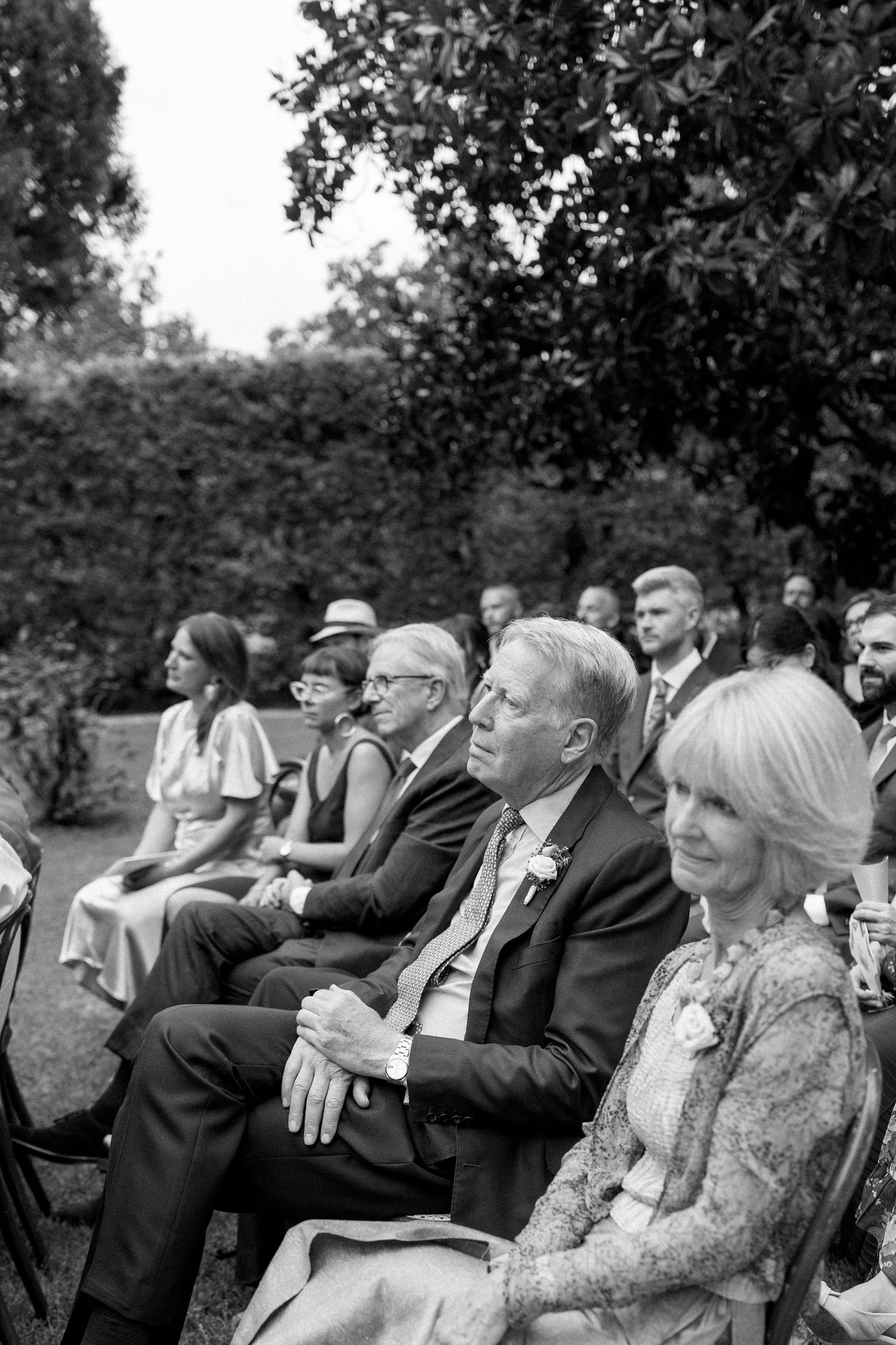 Wedding guests watch attentively during an outdoor ceremony seated on wooden chairs.