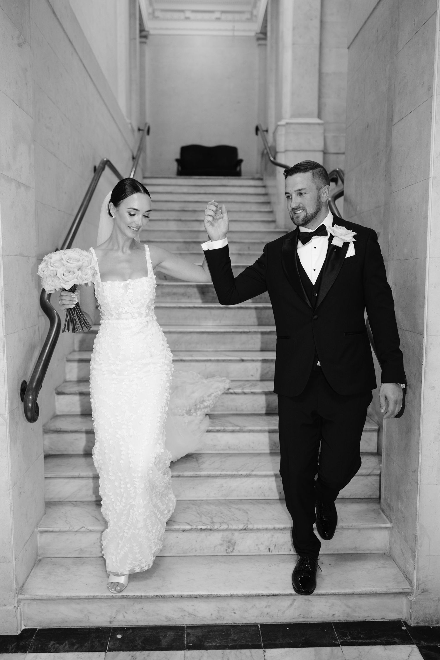 Playful wedding couple poses on elegant white marble staircase in classic black and white style.