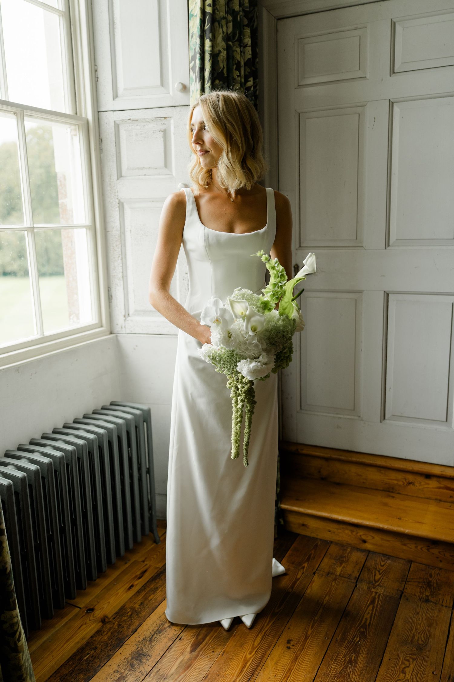 Bride in elegant white satin gown holding green and white bouquet by window.