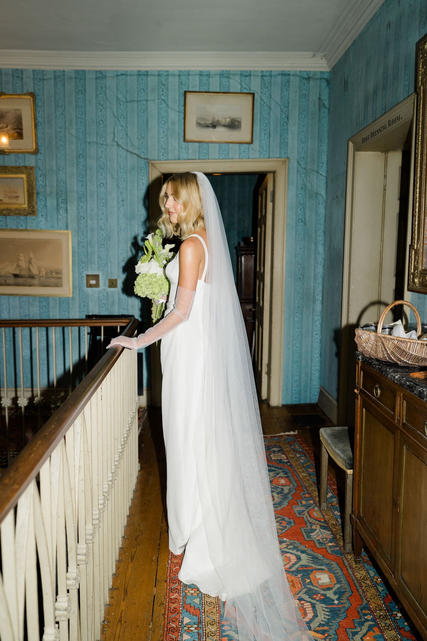 Elegant bride in white gown and veil holding green bouquet on staircase landing in blue hallway.