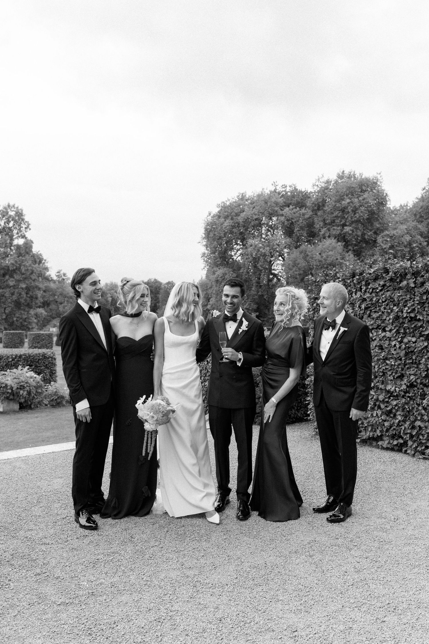 Black and white photo of a wedding couple posing with family in formal attire outdoors.