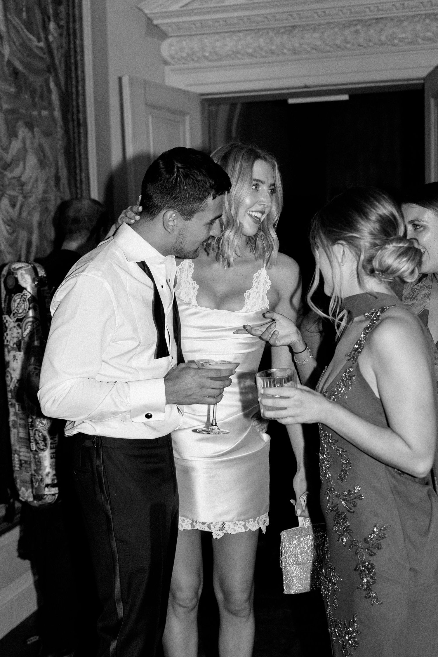 Bride in white mini dress laughs with guests holding cocktails at wedding reception.