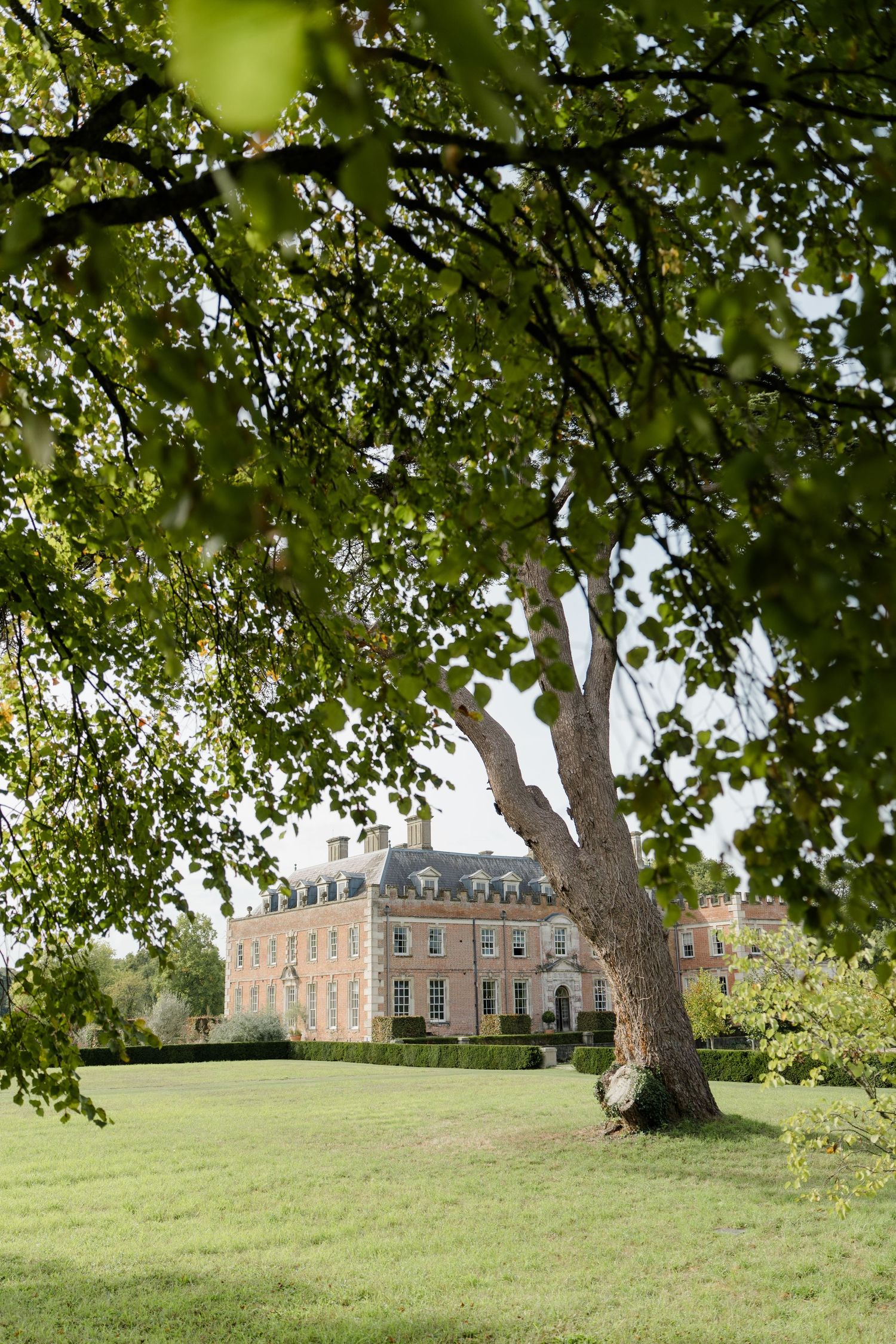 A grand pink Georgian manor house framed by lush green tree branches on a sunny day.