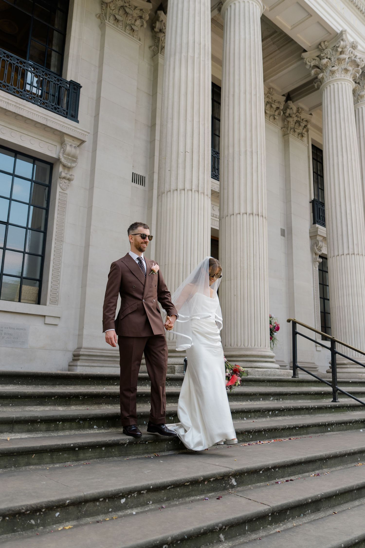 Bride and groom hold hands descending stone steps of neoclassical building with tall white columns.