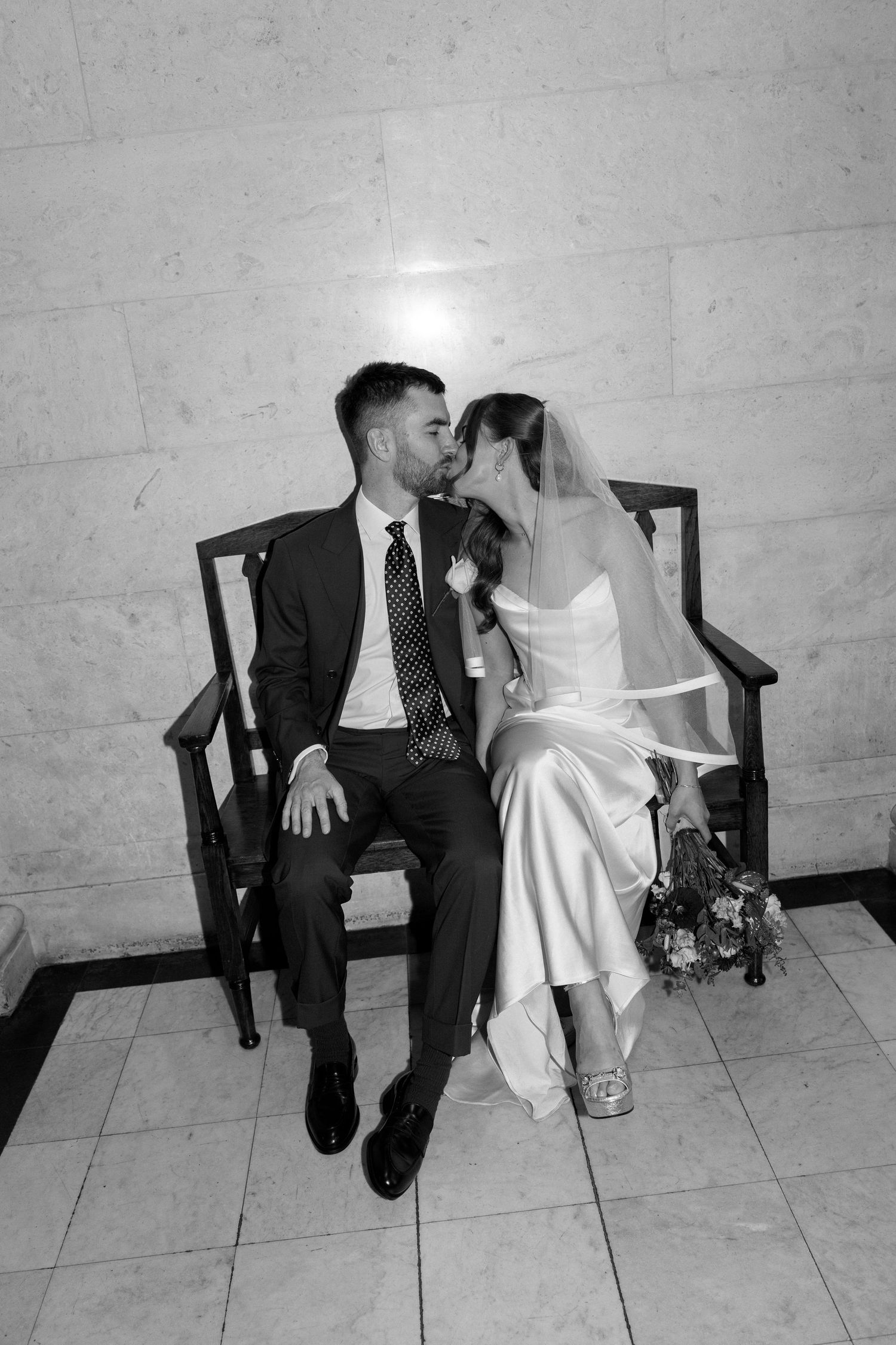 Black and white photo of bride and groom sharing a kiss while seated on a wooden bench.
