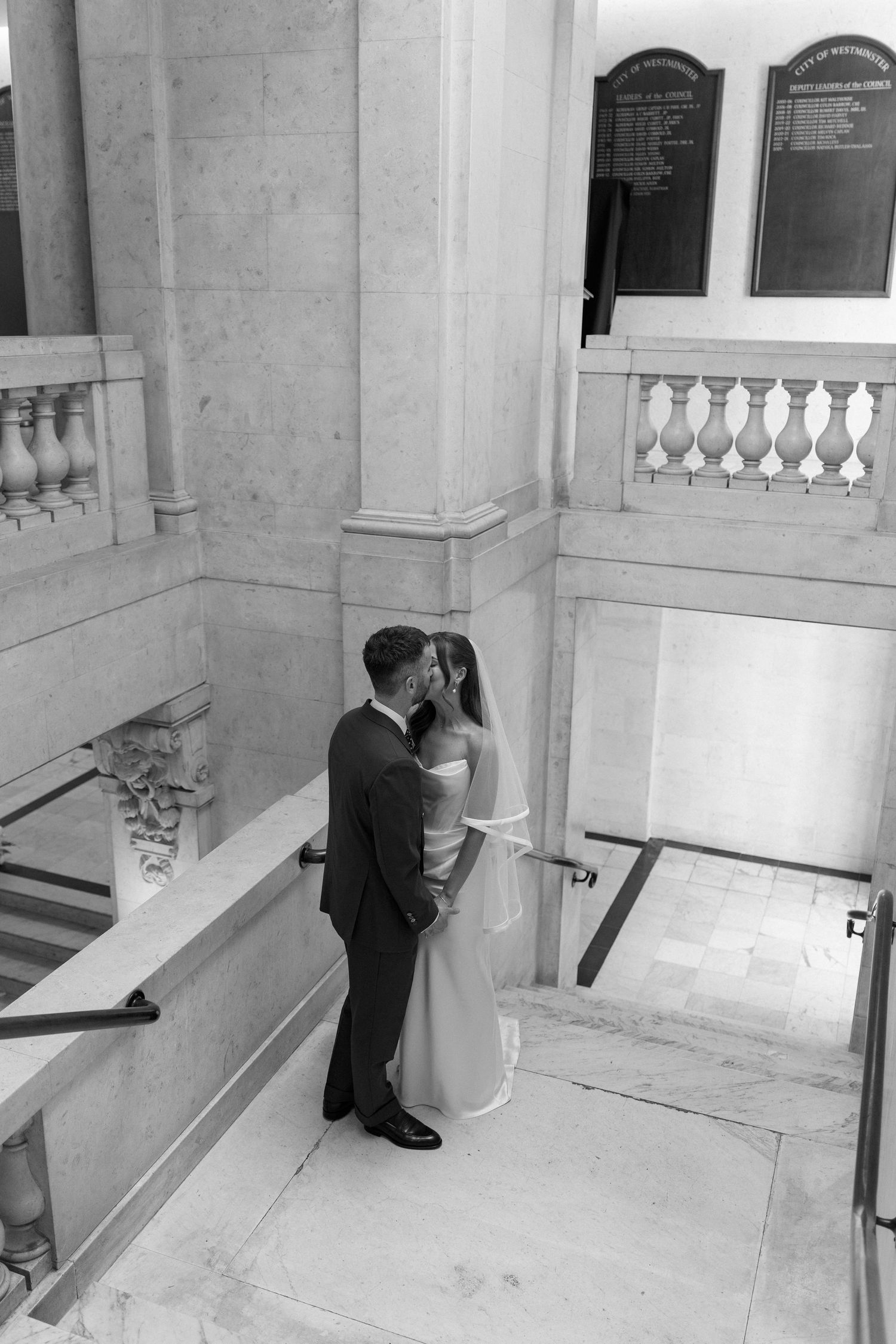 Black and white photo of newlyweds sharing a kiss on a grand marble staircase.